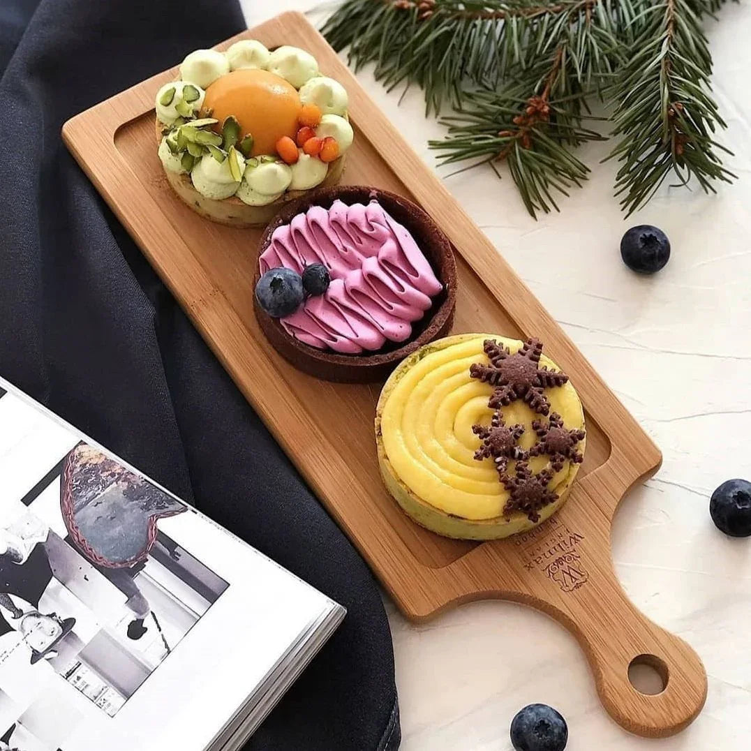 Three colorful gourmet tarts with decorative toppings on a wooden serving board with blueberries and pine branches nearby