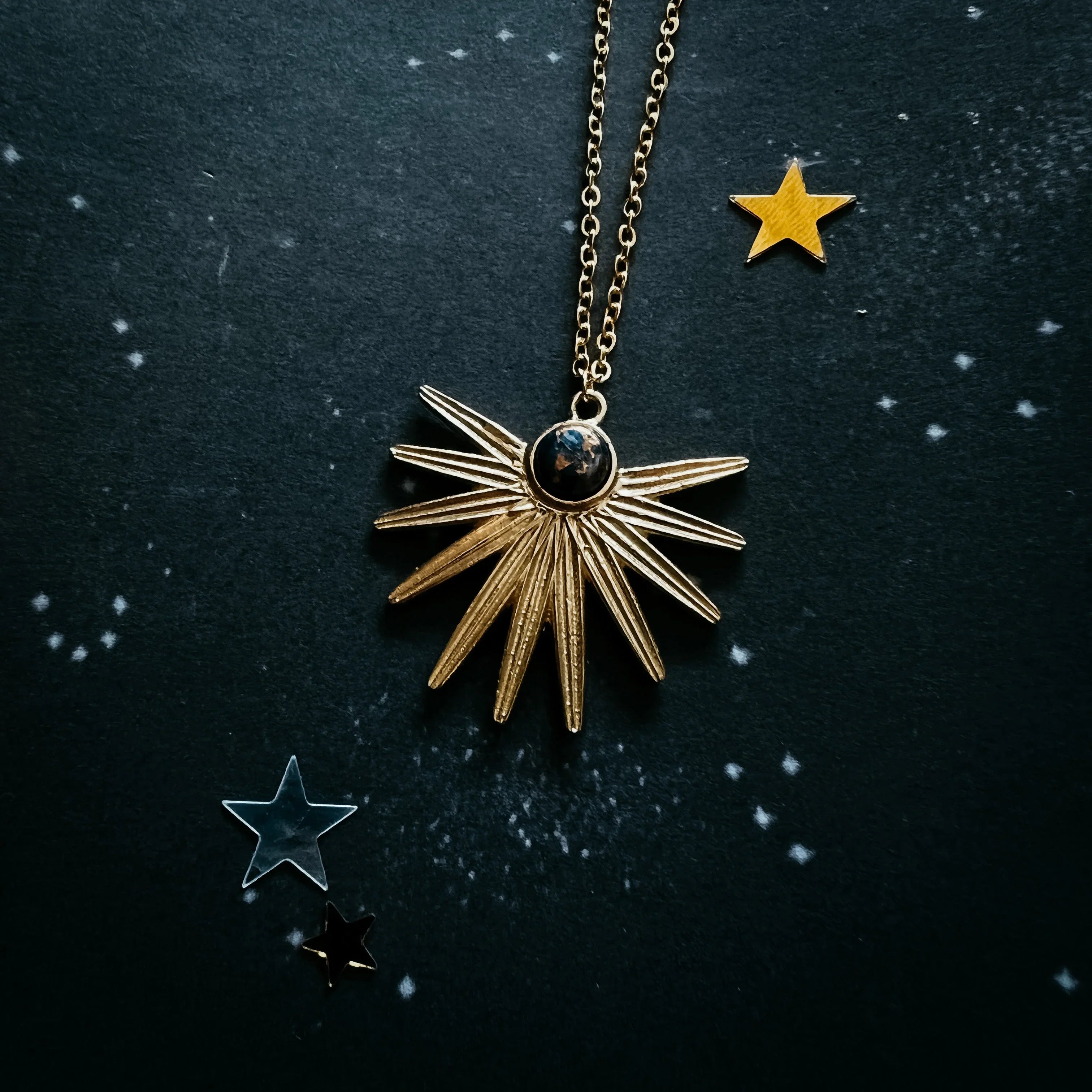 Gold sunburst pendant necklace with black stone on black starry background with decorative stars