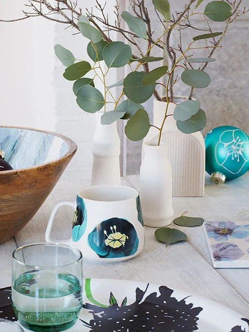 White ceramic mug with blue floral design on a table with a green glass, wooden bowl, abstract plate, eucalyptus branches in vases, and decorative ball