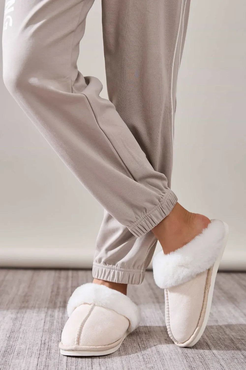 Close-up of person wearing beige jogger pants and white fluffy fleece slippers on wooden floor