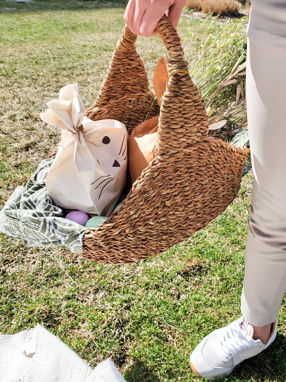 Hand holding a woven basket with Easter gifts, colorful eggs, and spring greenery on grass