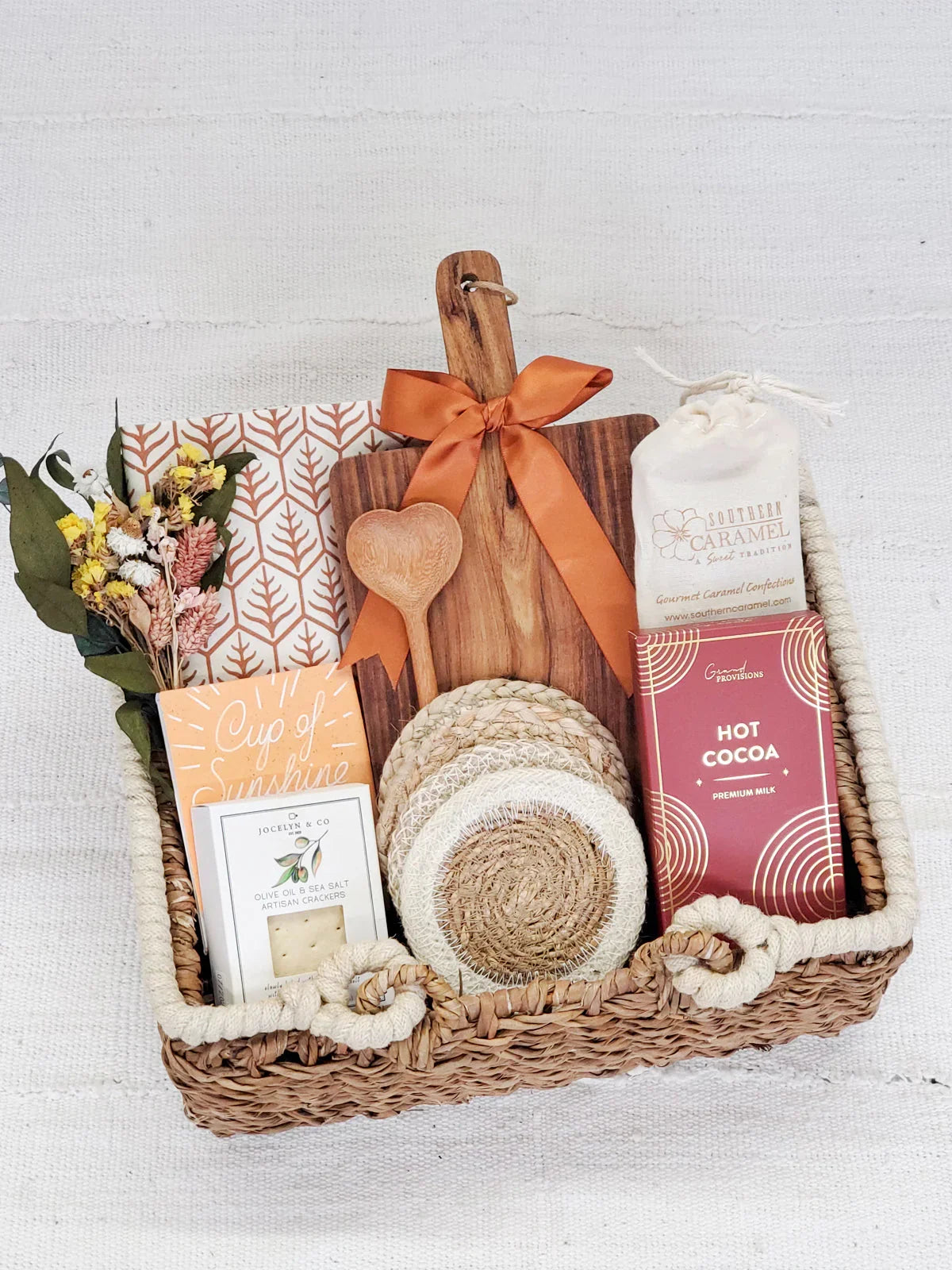 Wicker gift basket with hot cocoa, caramel, crackers, wooden cutting board, heart spoon, coasters, and flowers