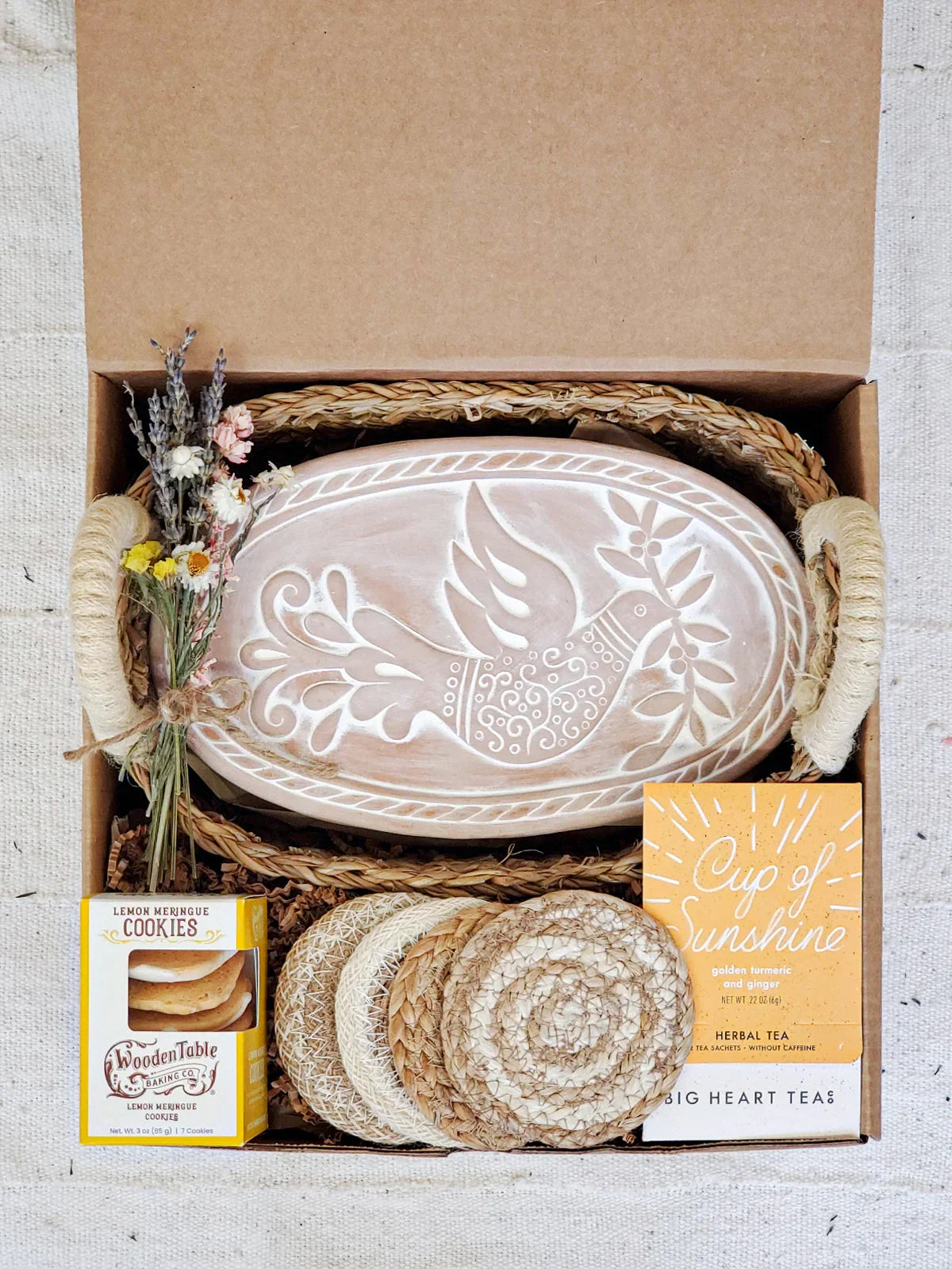 Gift box with oval wooden bread warmer featuring bird design, dried flowers, tea, cookies, and woven coasters