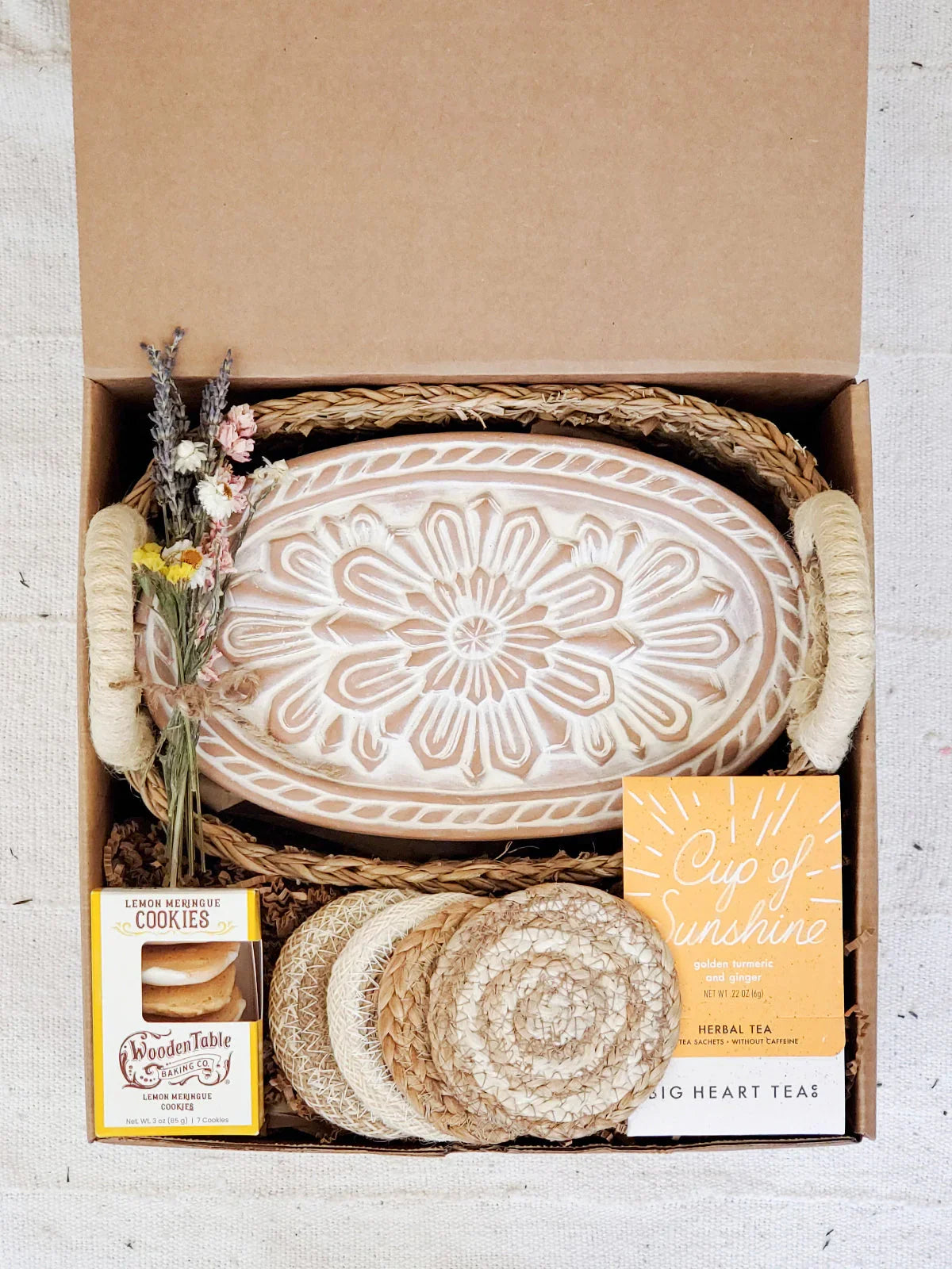 Gift box with oval ceramic bread warmer, dried flowers, coasters, lemon cookies, and herbal tea