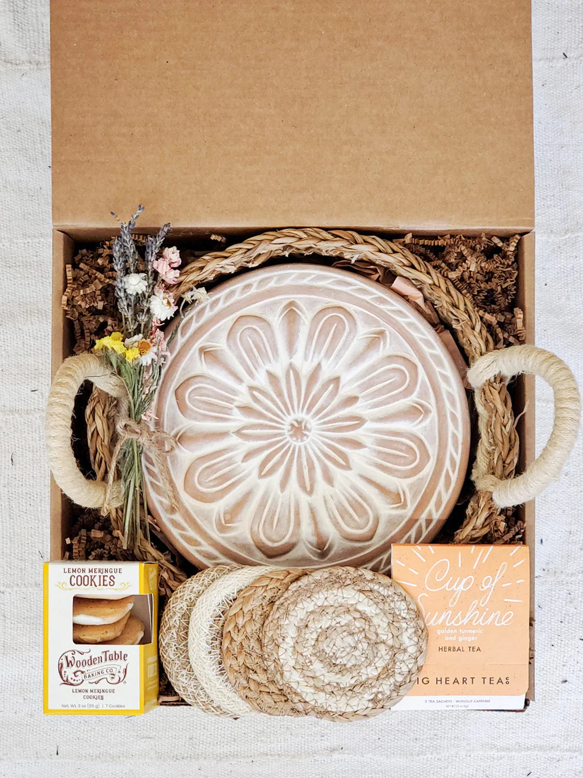 Gift box containing ceramic bread warmer with floral design, lemon cookies, herbal tea, coasters, and dried flowers