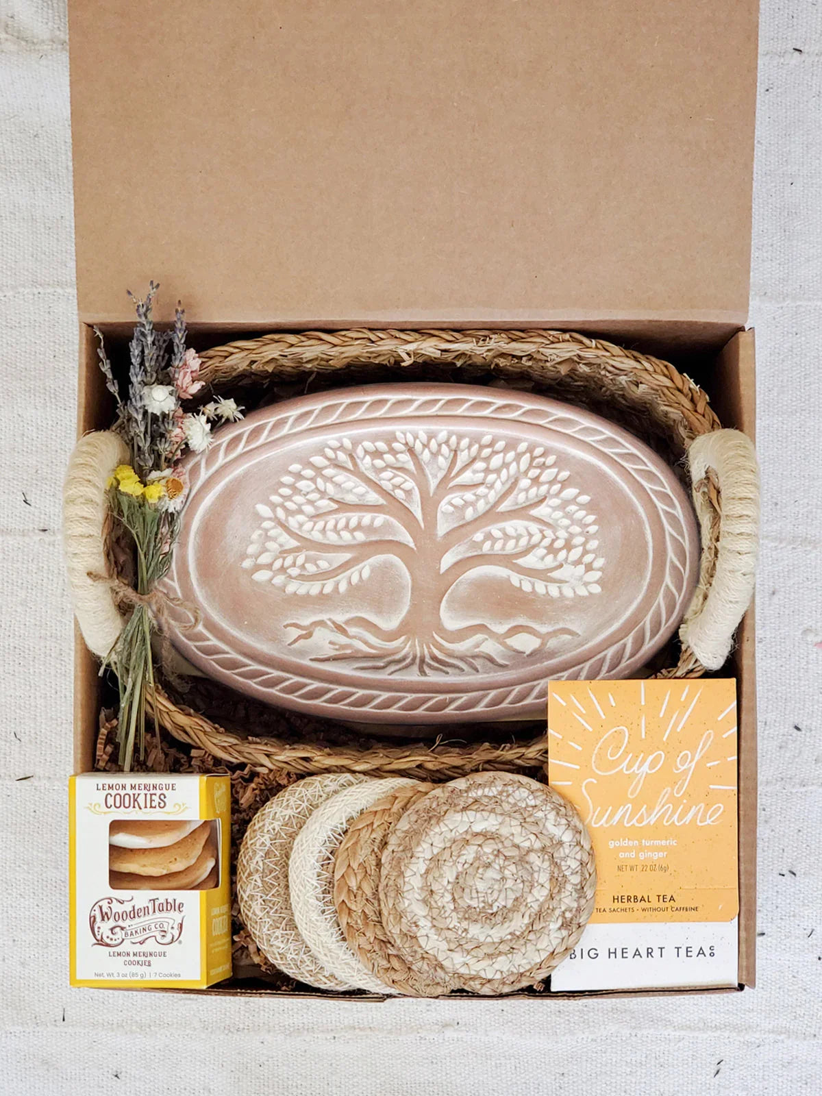 Gift box with ceramic bread warmer featuring tree design, herbal tea, lemon meringue cookies, coasters, and dried flowers