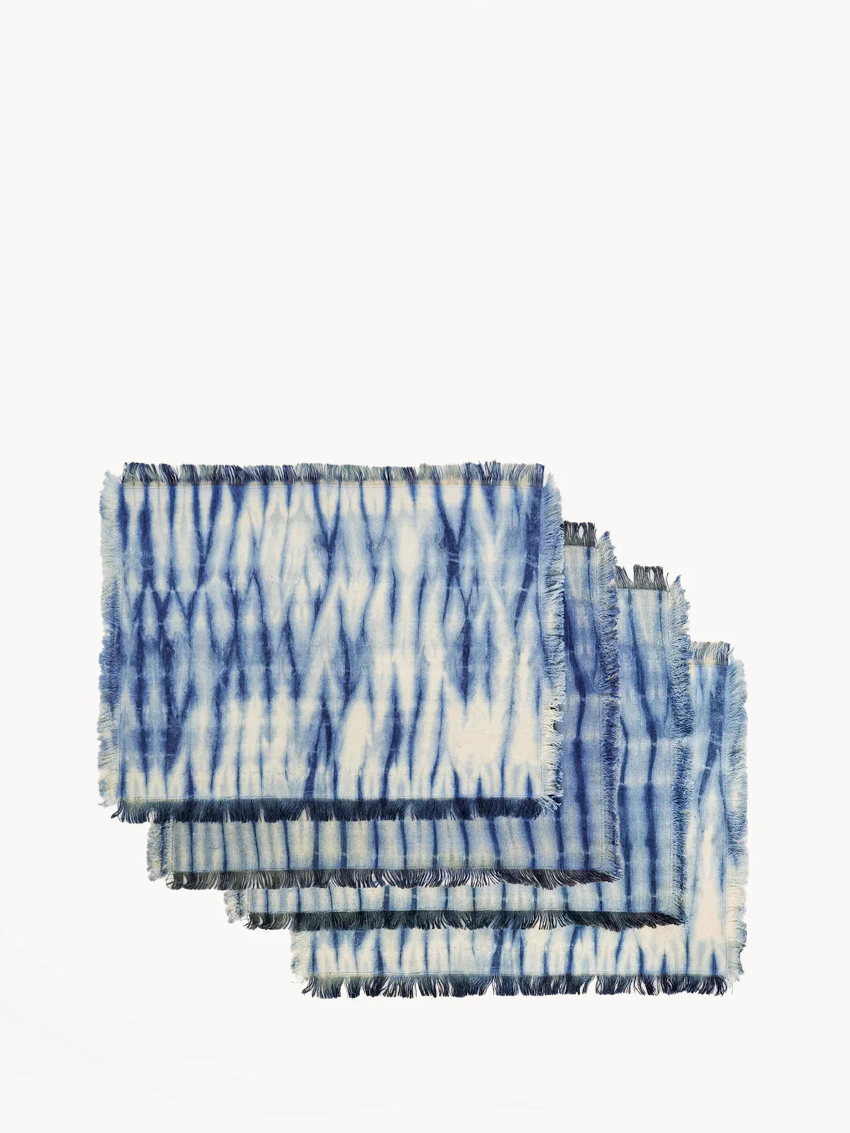 Four blue and white tie-dye cotton placemats with fringed edges stacked on white background