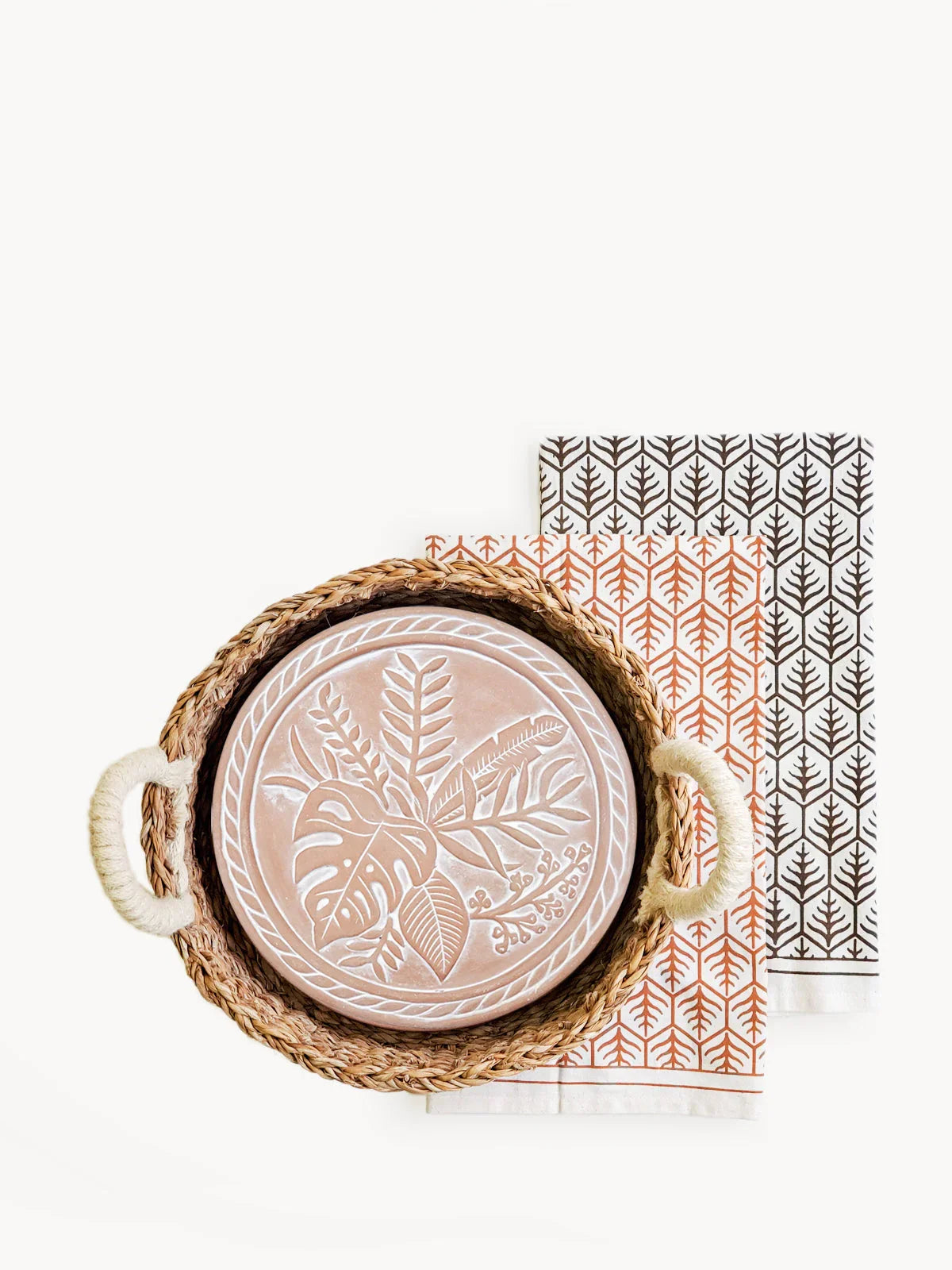 Round woven basket with rope handles holding a decorative terracotta plate with leaf patterns, on two patterned tea towels