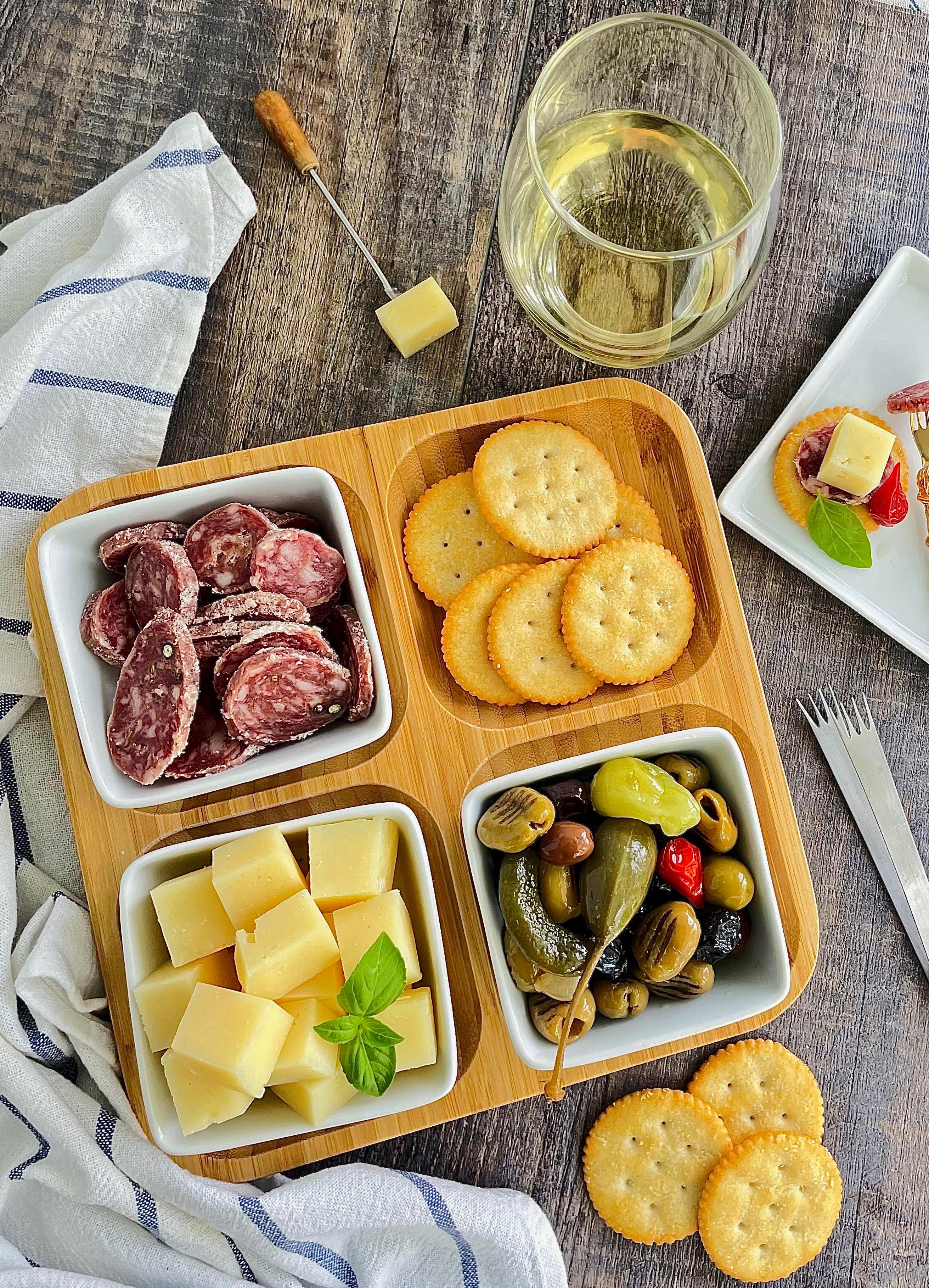 Charcuterie board with salami, cheese cubes, crackers, olives, and a glass of white wine on wood table