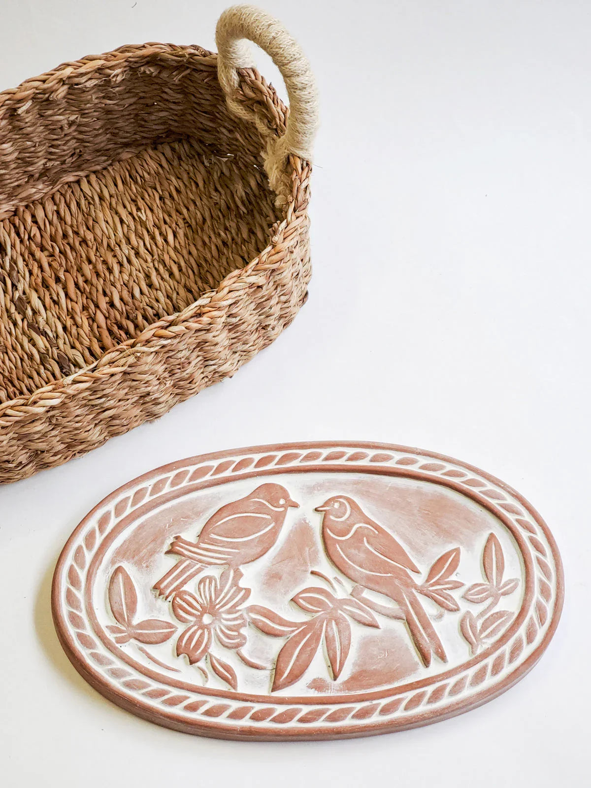 Oval terracotta bread warmer with carved lovebird and floral design beside woven basket