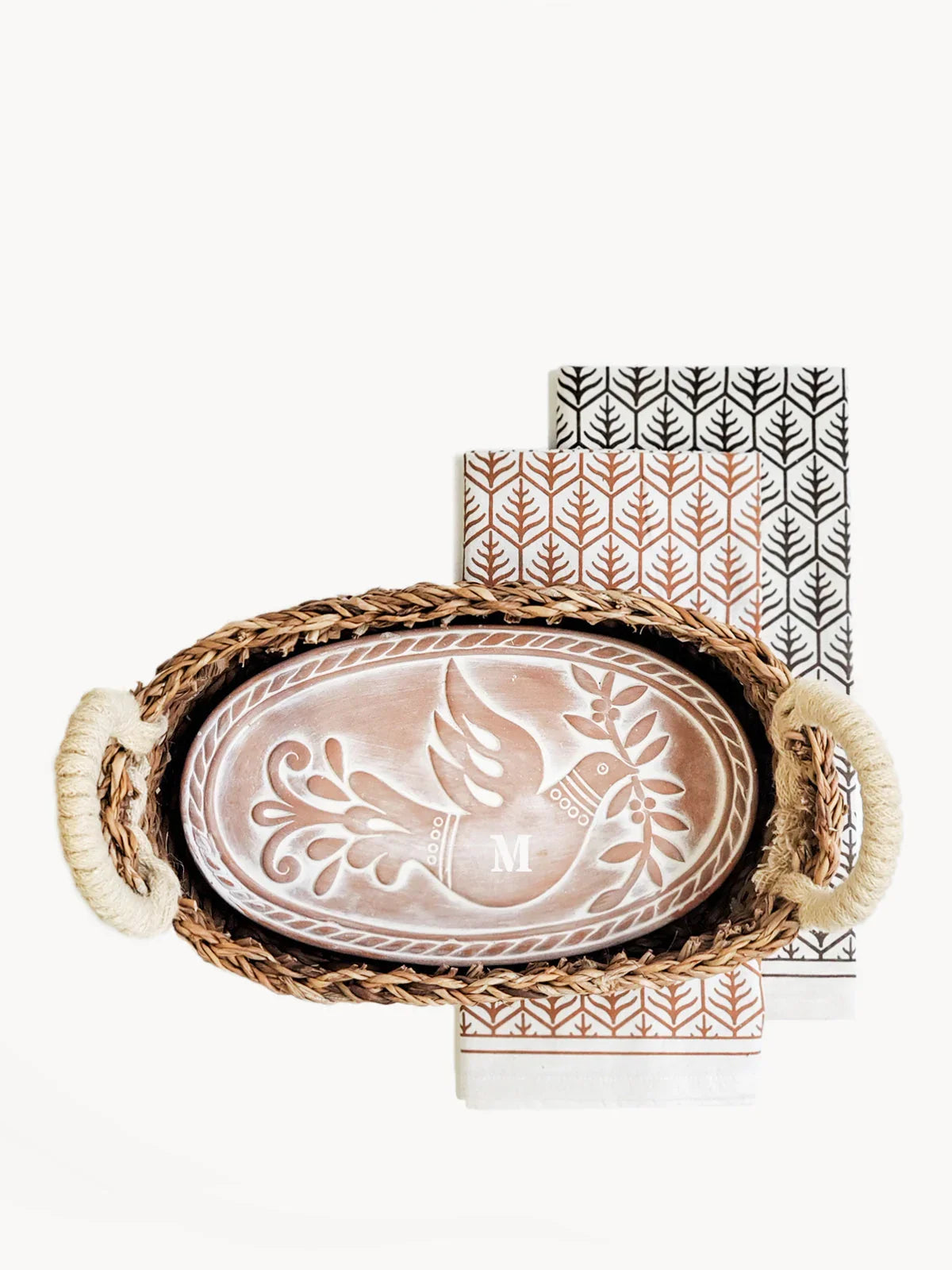 Oval ceramic bread warmer with embossed bird design and monogram M in woven basket with patterned cloth napkins