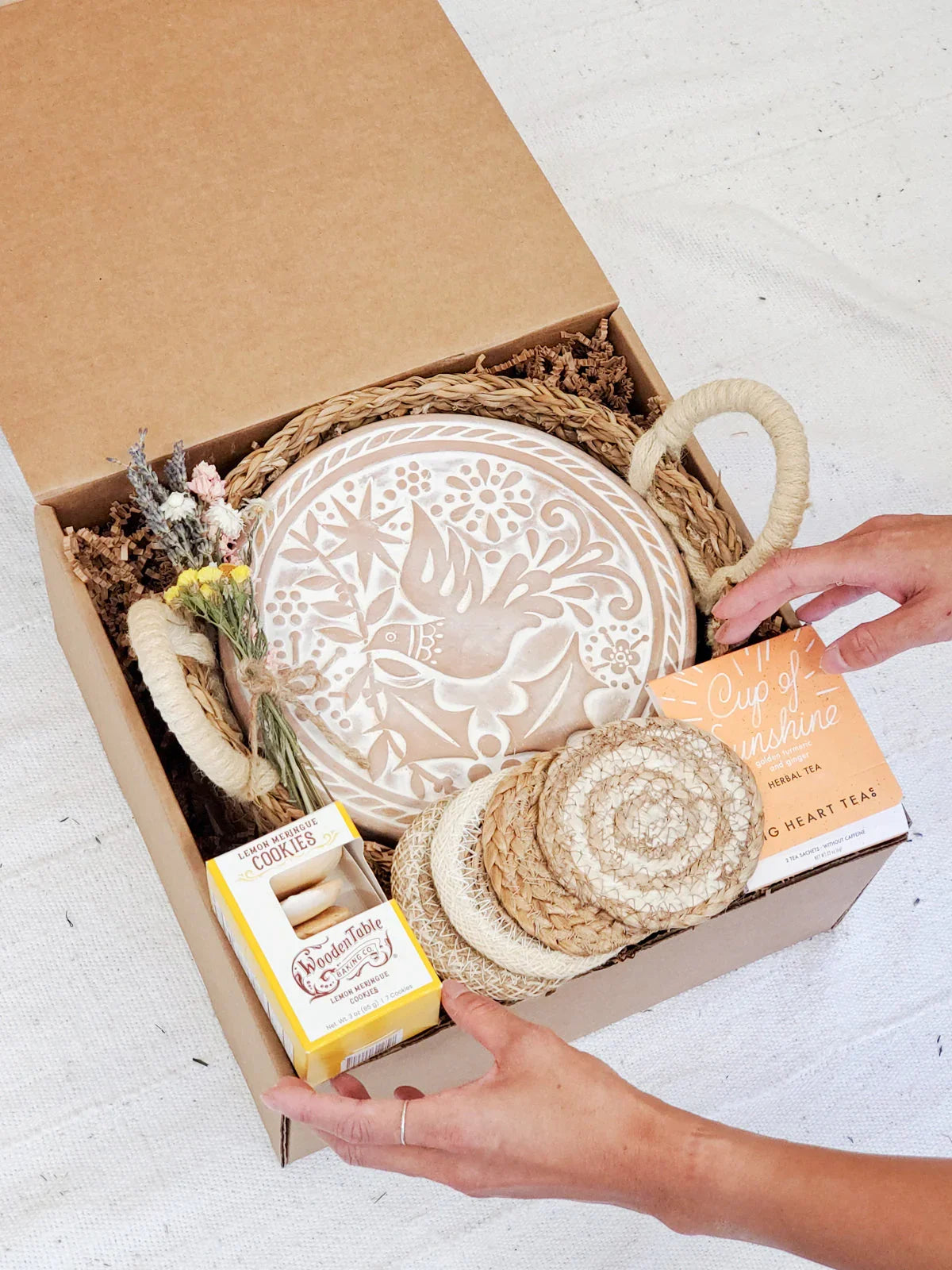 Gift box with carved bread warmer, woven coasters, dried flowers, lemon meringue cookies, and herbal tea