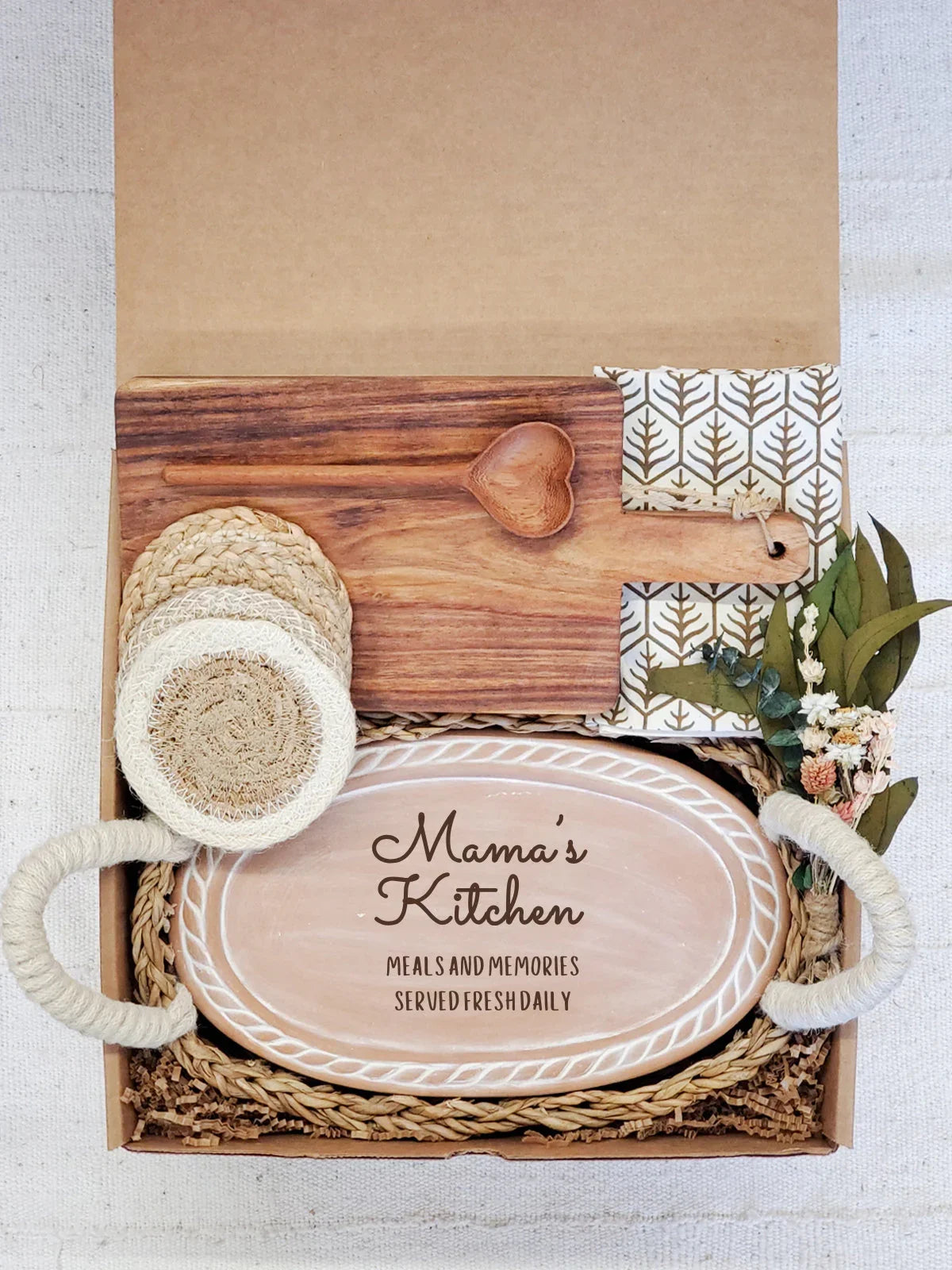 Gift box with decorative wooden cutting board, heart-shaped spoon, patterned napkin, woven coasters, and oval serving tray inscribed Mama's Kitchen