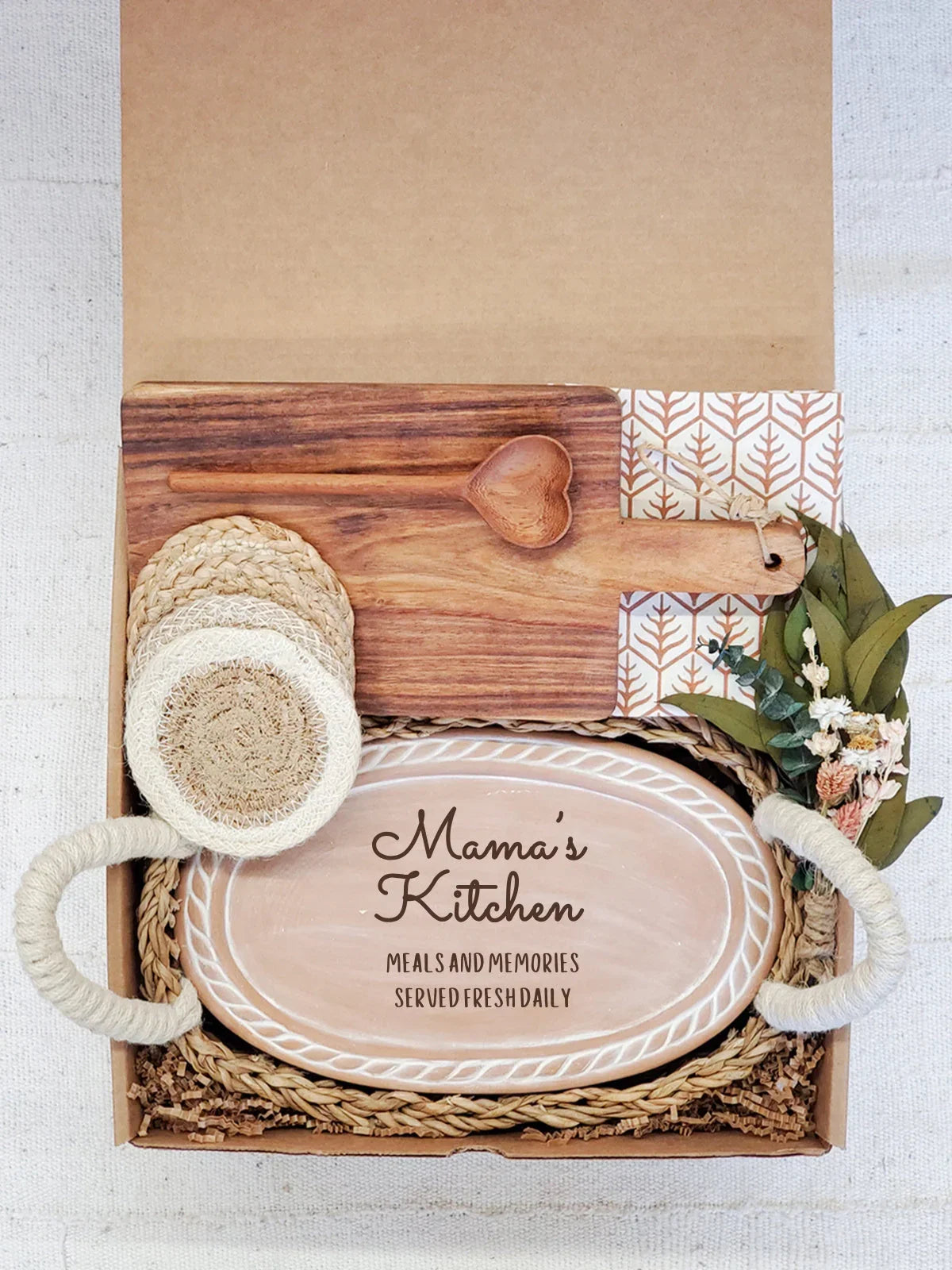 Gift box with a wooden heart spoon, cutting board, woven coasters, and an oval tray saying 'Mama's Kitchen'