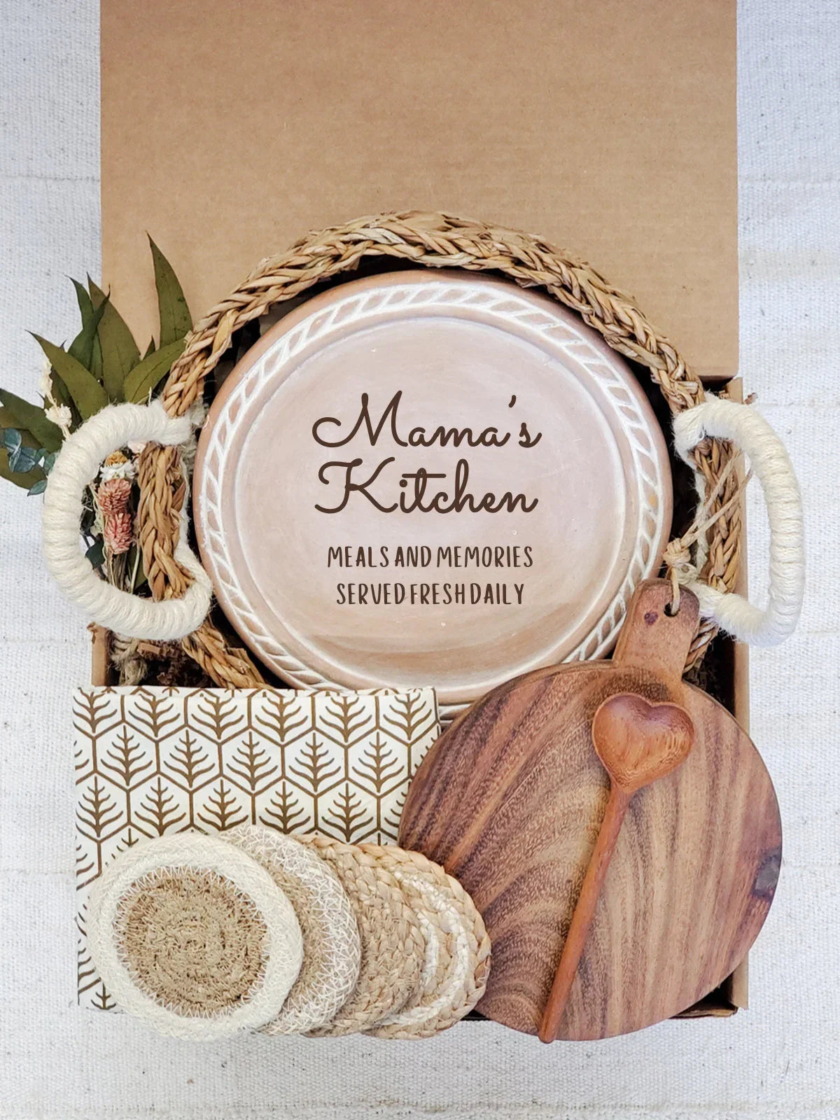 Gift box with woven basket, ceramic plate saying Mama's Kitchen, wooden cutting board, heart-shaped spoon, coasters, and patterned napkin