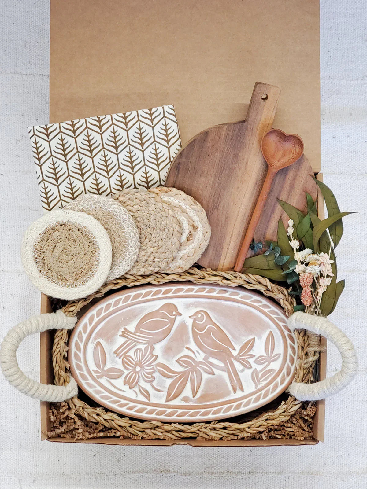 Gift box with rustic kitchen items including oval bread warmer with bird design, wooden cutting board, coasters, and dried flowers