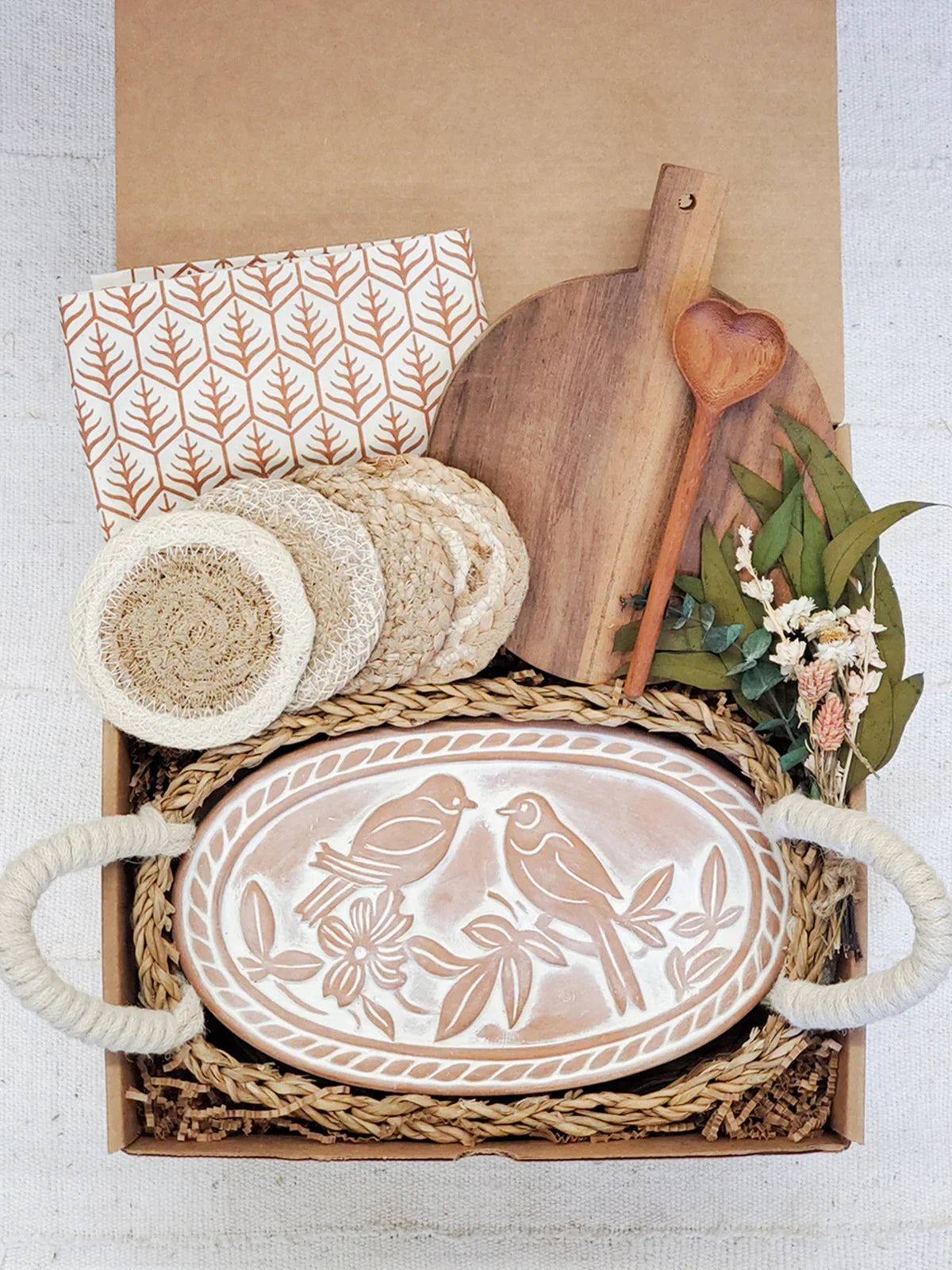Decorative gift box with a bird-themed ceramic bread warmer, woven coasters, wooden cutting board, heart-shaped wooden spoon, and dried flowers