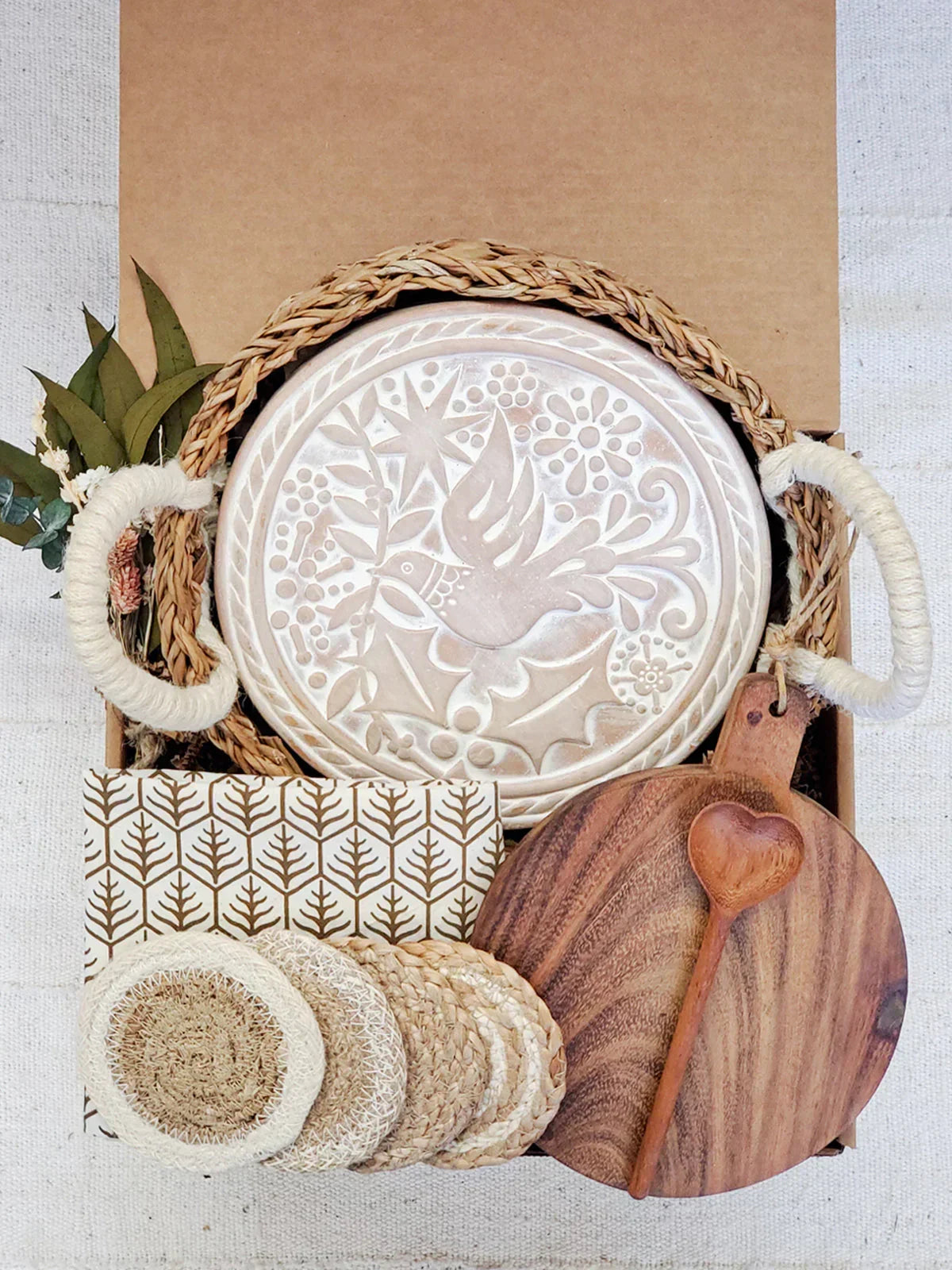 Rustic gift box with round bread warmer featuring bird design, wooden cutting board, heart-shaped spoon, coasters, and decorative leaves
