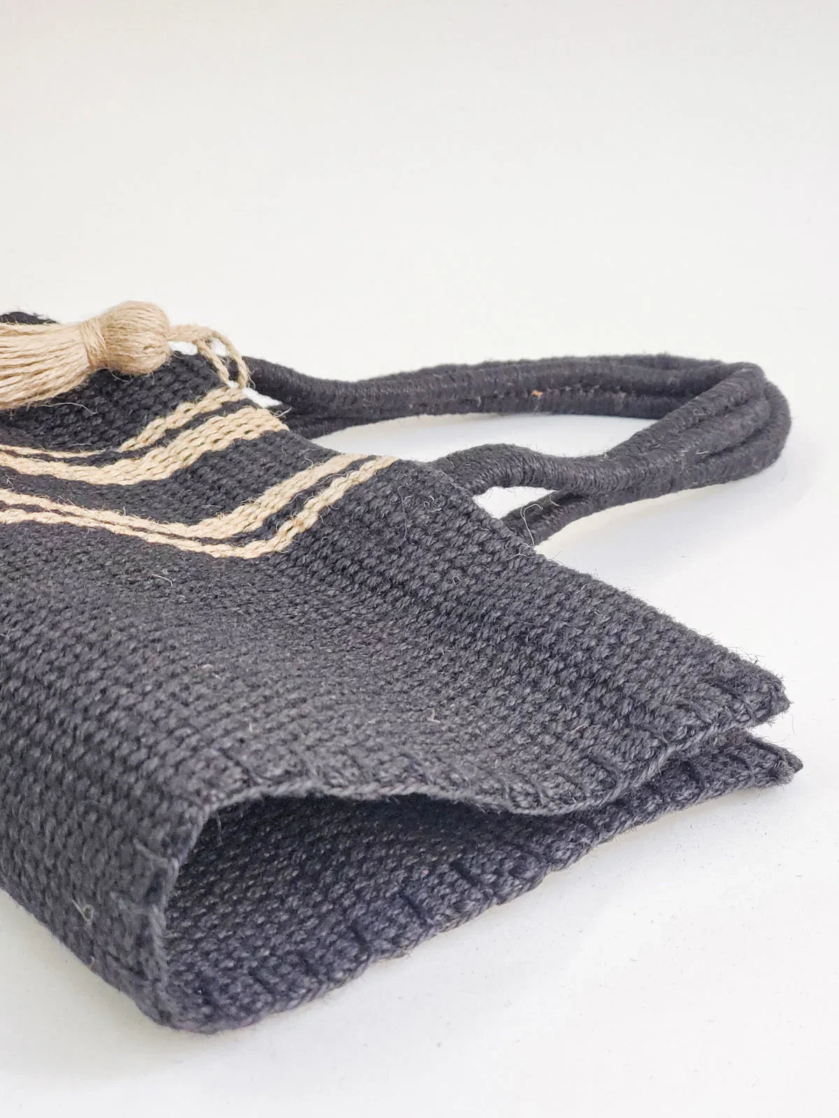 Handwoven black jute tote bag with beige stripes and tassel detail on white background