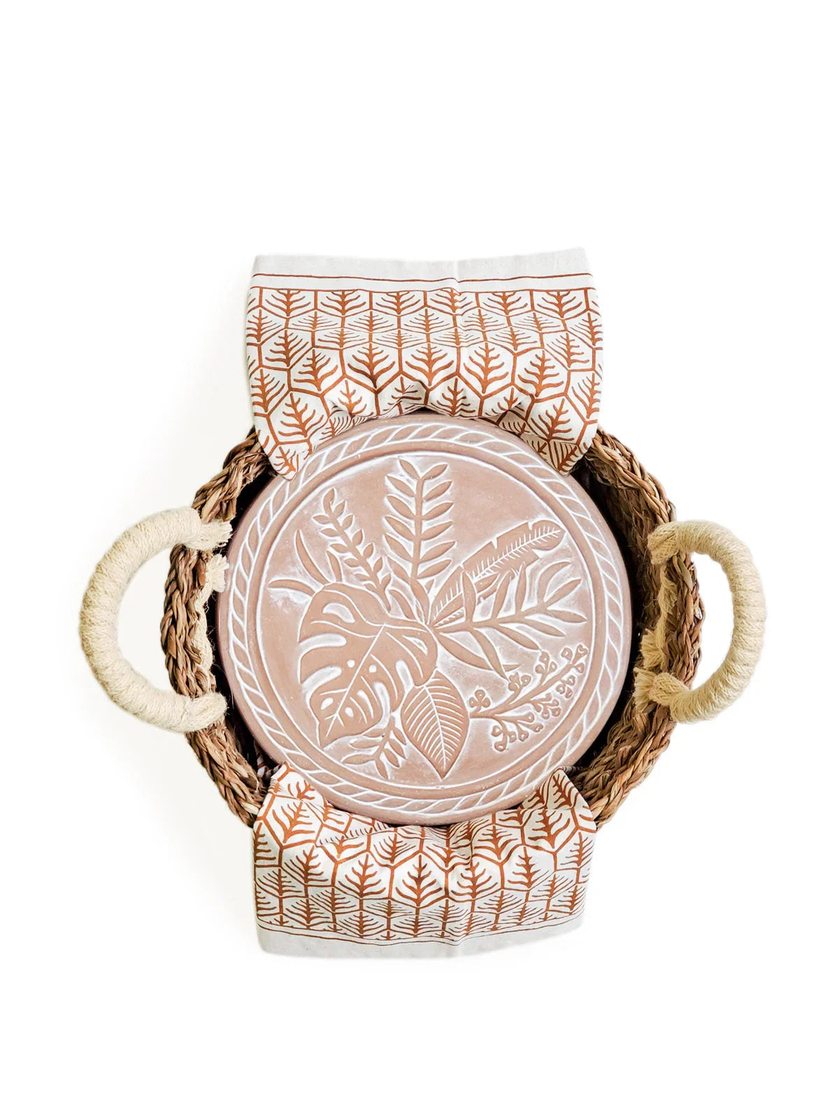 Woven round basket with rope handles holding a round terracotta plate with leaf designs and a patterned tea towel