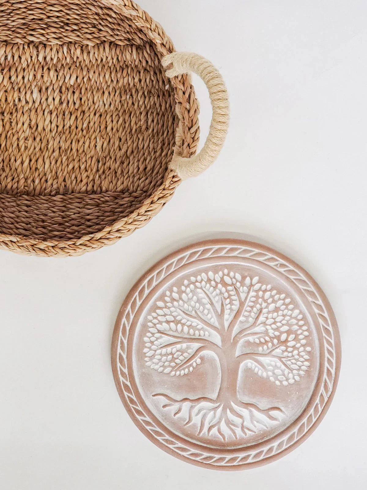 Round terracotta bread warmer with tree of life design next to woven basket on white surface