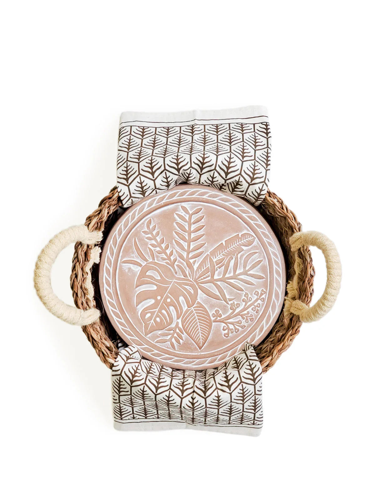 Woven basket with rope handles holding a round ceramic plate with embossed leaves and patterned tea towel