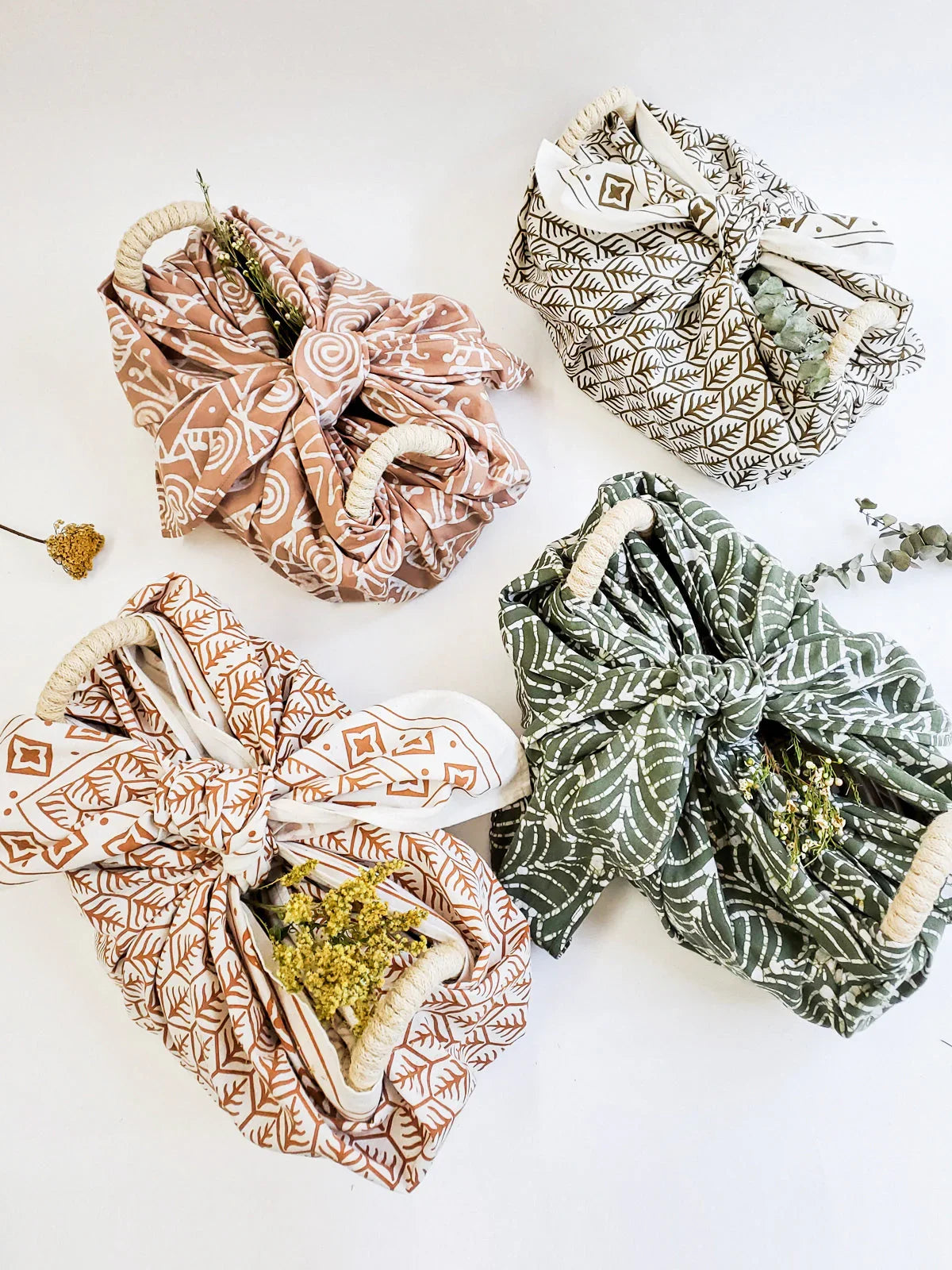 Four patterned fabric bread warmer baskets with rope handles, tied with bows and decorated with dried flowers on white background
