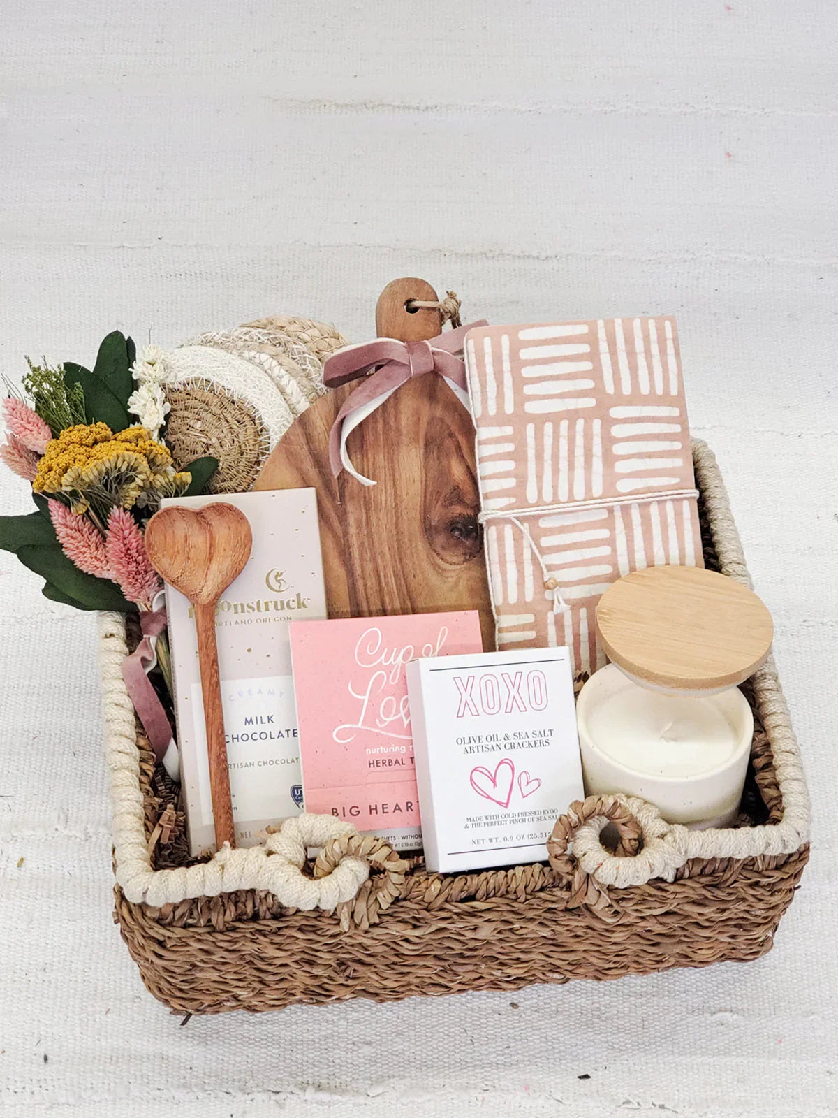 Rustic gift basket with wooden spoon, chocolate, herbal tea, artisan crackers, cutting board, patterned cloth, candle, and dried flowers