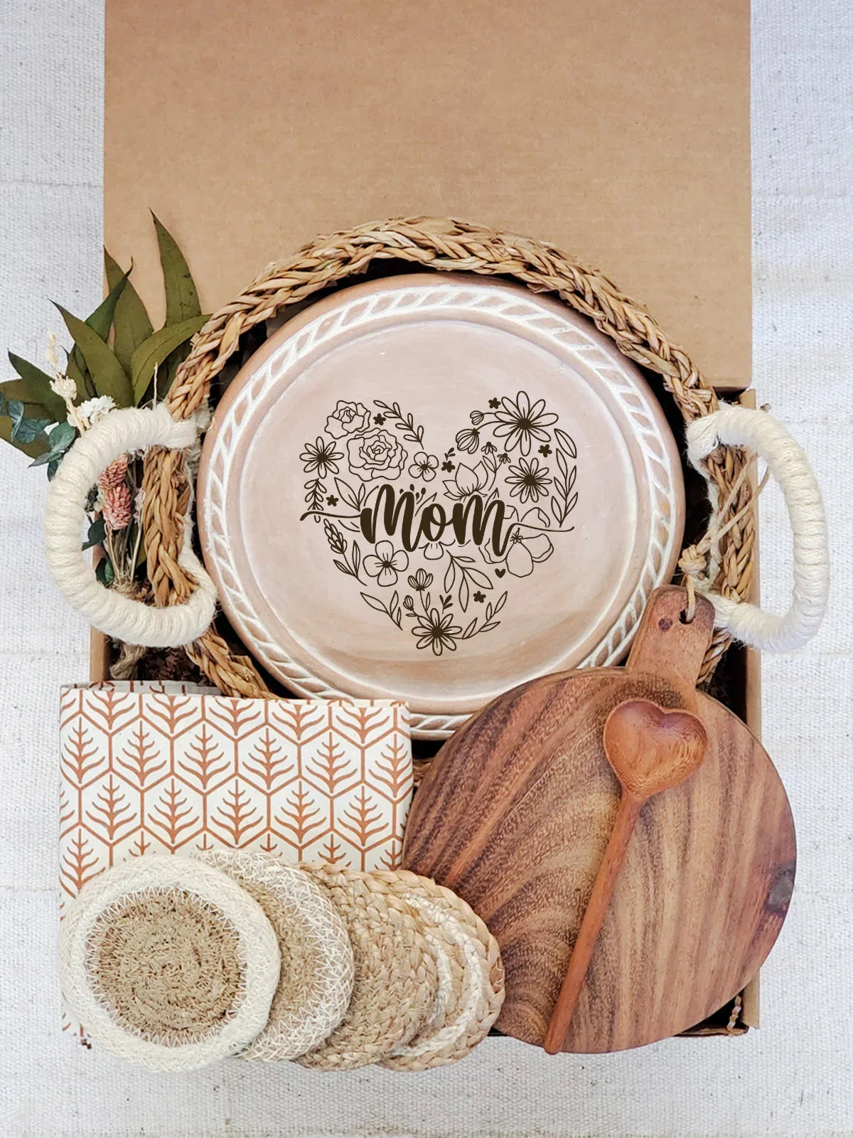 Gift box with round bread warmer featuring floral heart design and 'mom' lettering, wooden board, coasters, and floral decor