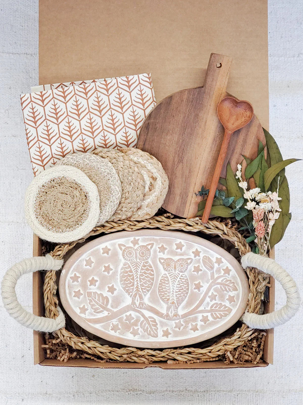 Gift box with oval ceramic bread warmer featuring owl design, wooden cutting board, heart spoon, coasters, and dried flowers