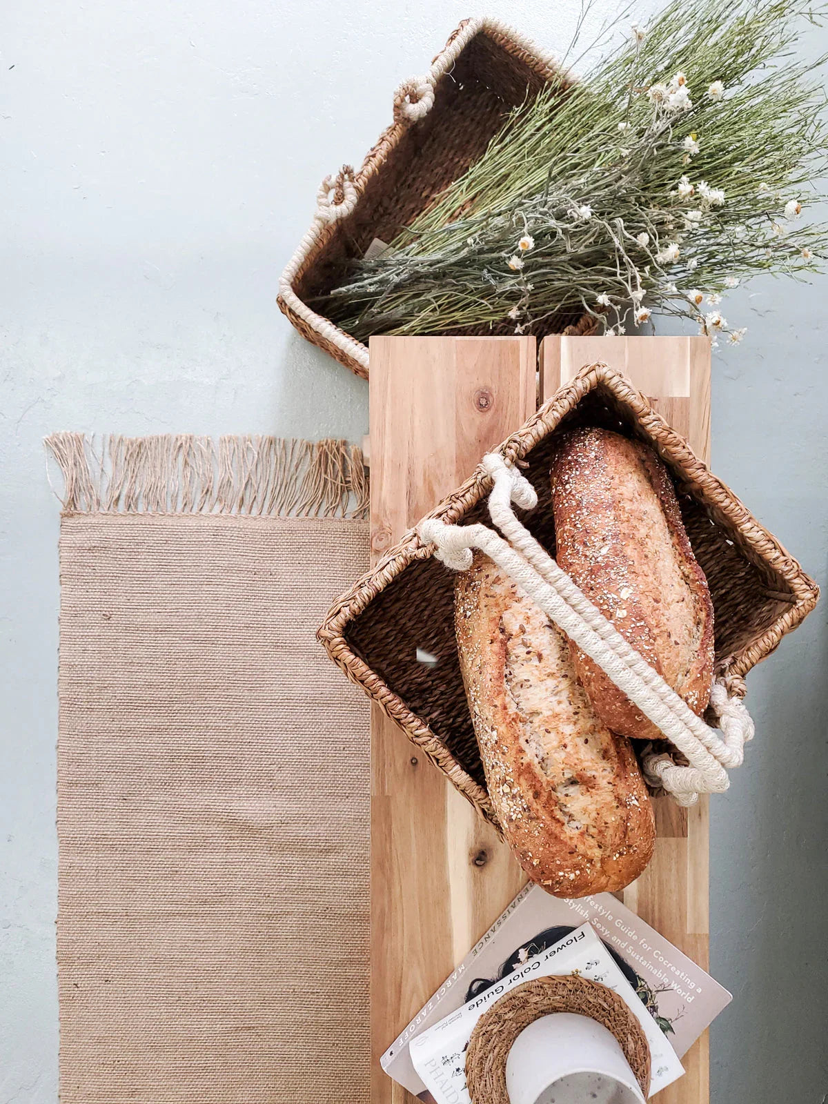 Woven storage basket with rope handles holding artisan bread, styled on wood with dried flowers