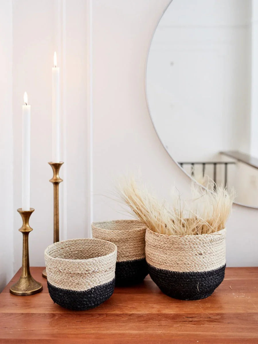Three woven storage bins with black and natural colors on wooden surface, two brass candlesticks with white candles, round wall mirror