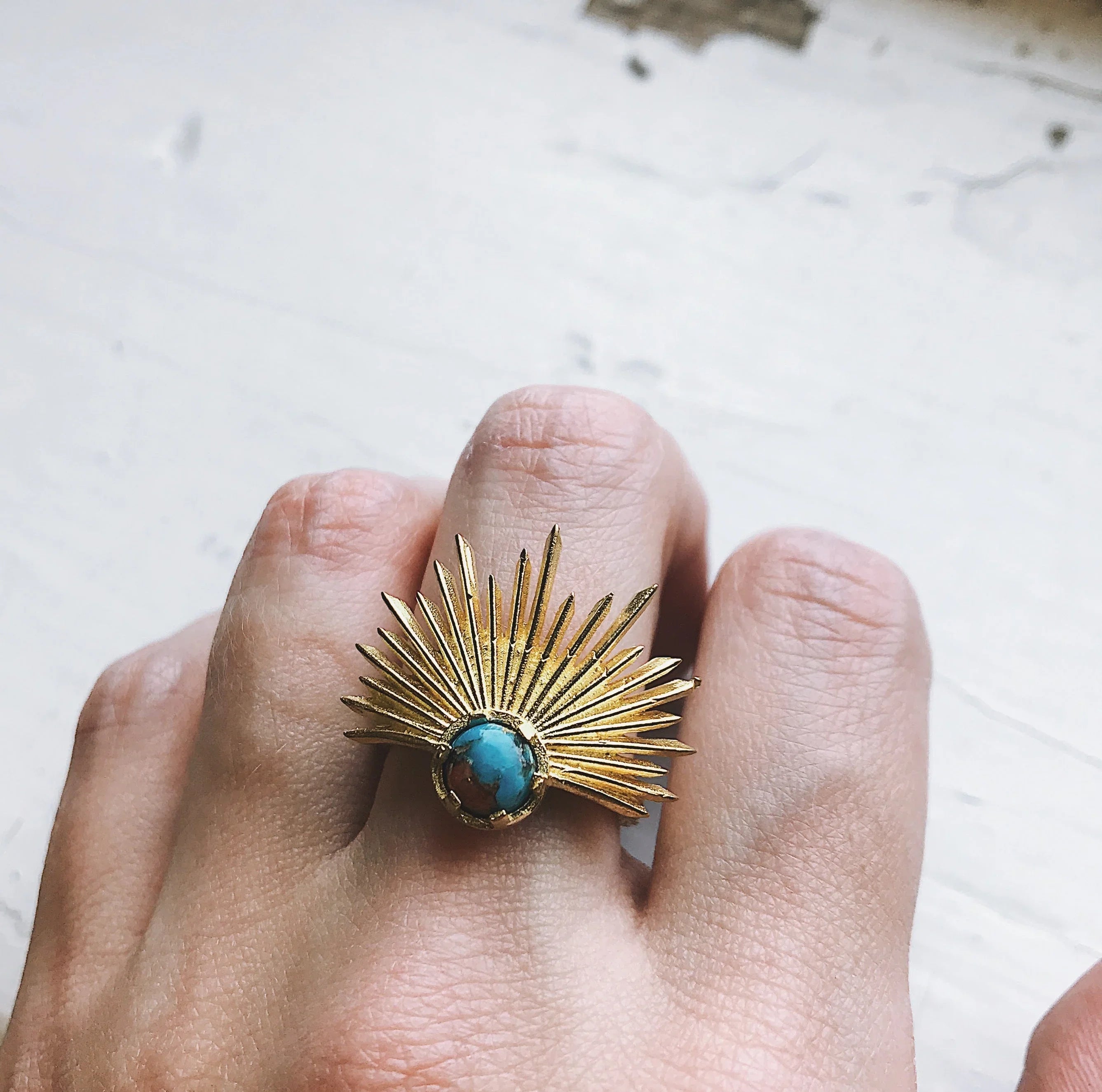 Gold sunburst ring with turquoise stone worn on finger against a light textured surface