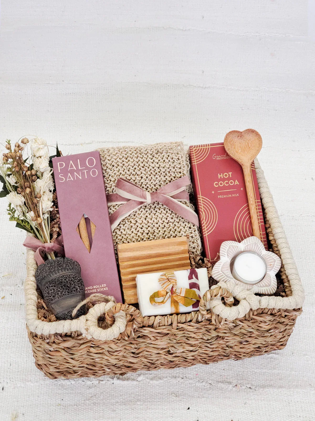 woven gift basket with Palo Santo incense, knitted cloth, hot cocoa, heart-shaped wooden spoon, soap, and flower candle holder
