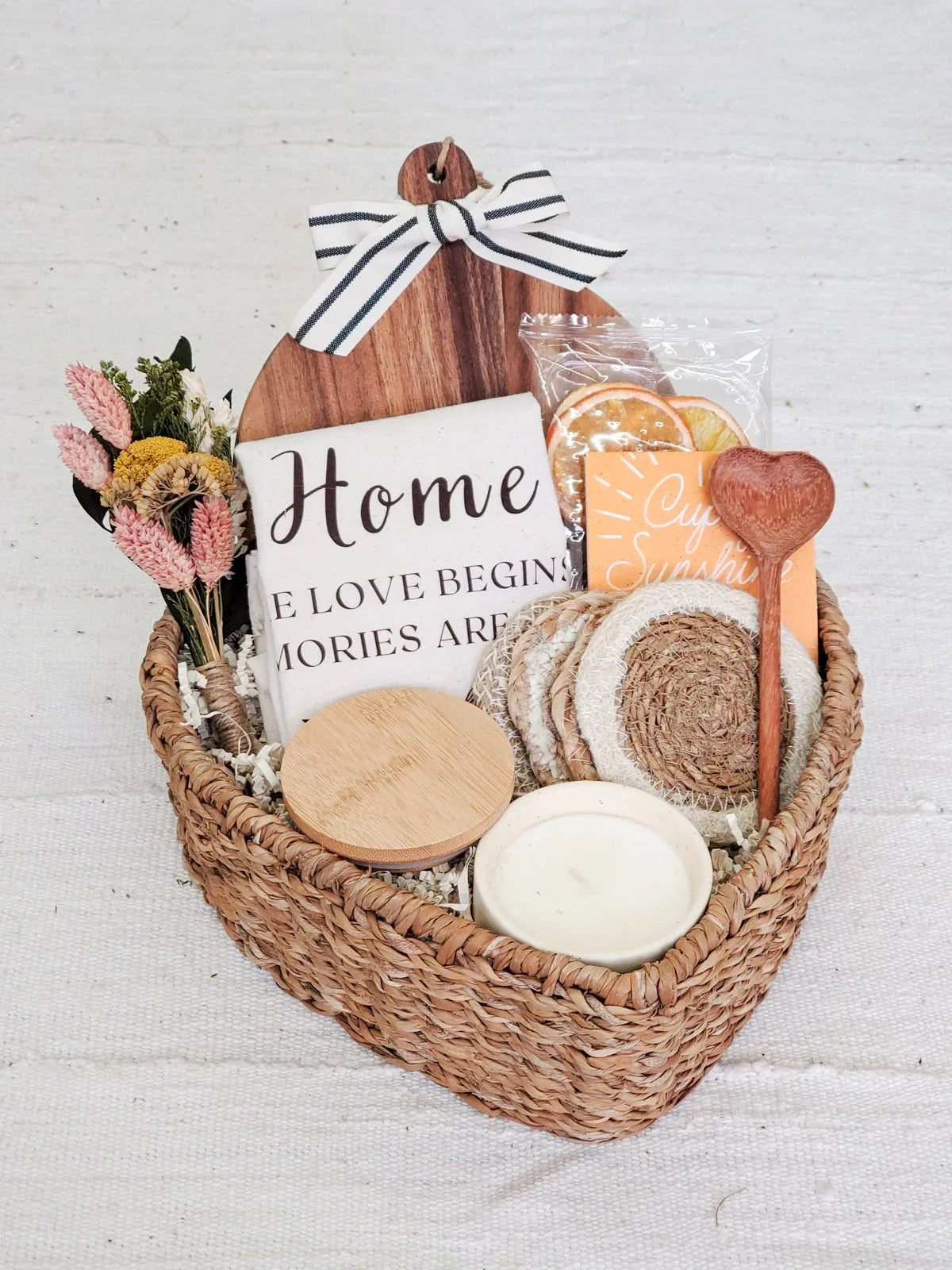 Heart-shaped woven gift basket with wooden cutting board, dried flowers, coasters, candle, and decorative items