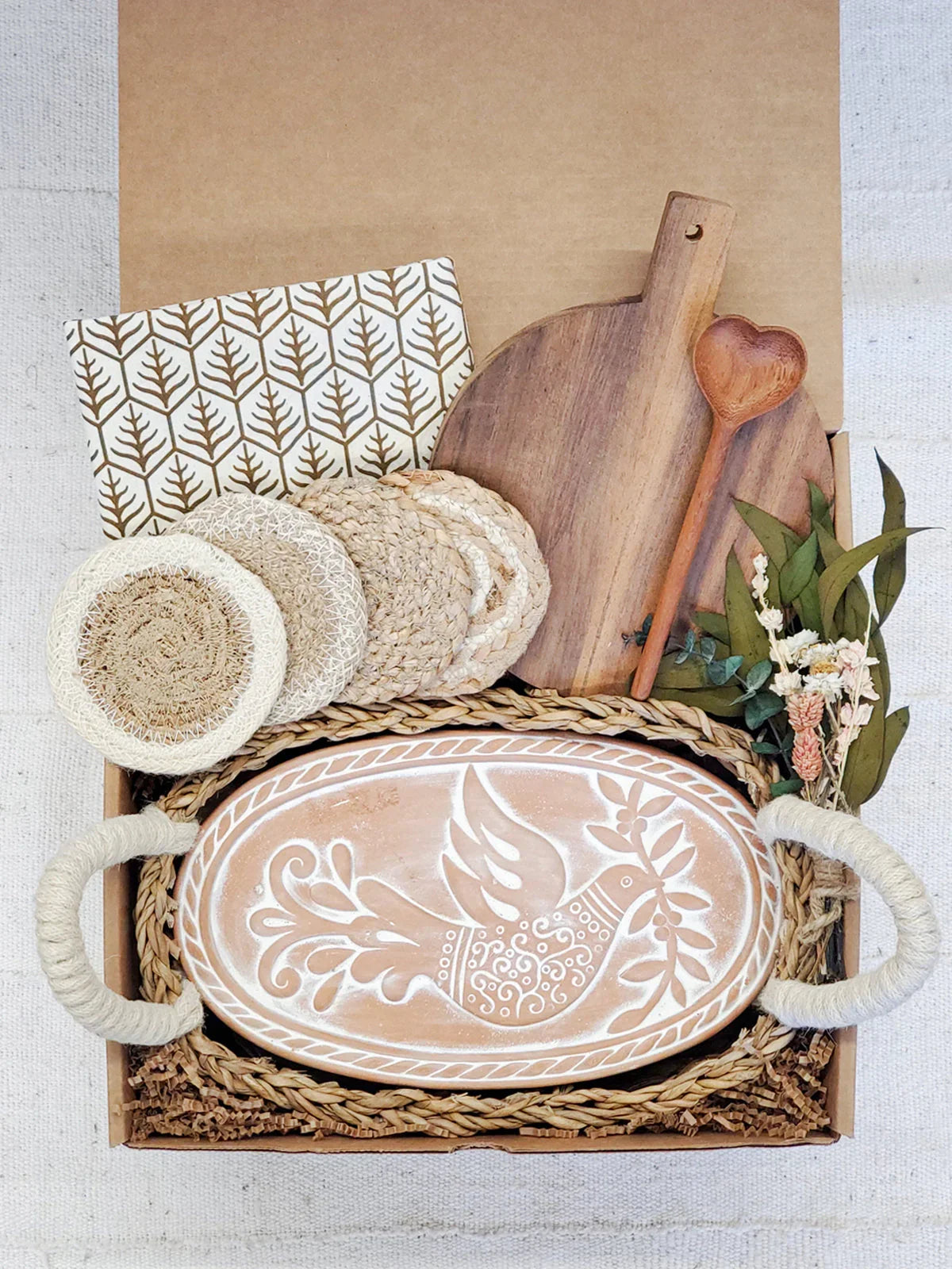 Gift box with oval ceramic bread warmer decorated with a bird, wooden cutting board, heart-shaped spoon, woven coasters, and dried flowers