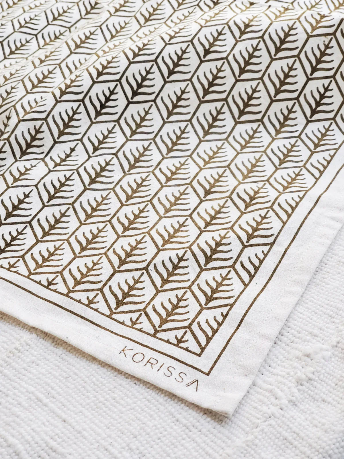 White tea towel with brown geometric leaf pattern and Korissa branding on textured white fabric
