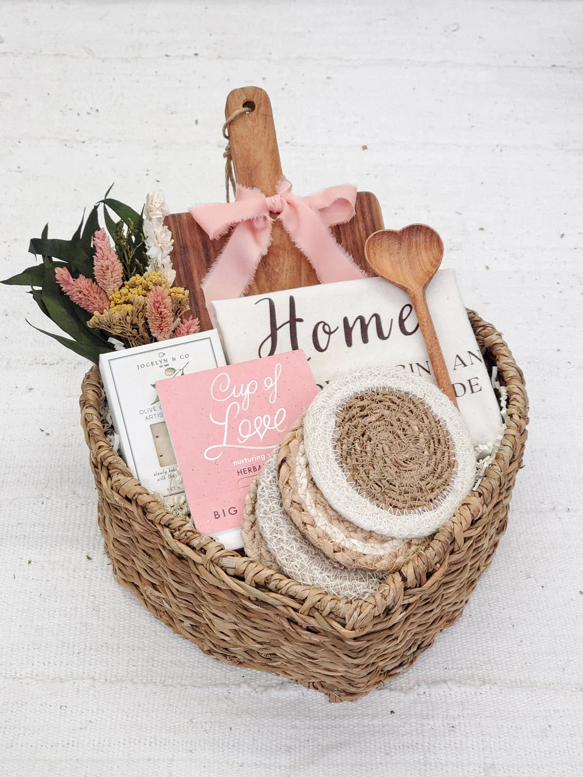 woven gift basket with wooden heart spoon, cutting board with pink bow, dried flowers, coasters, scented soap, and home decor towel