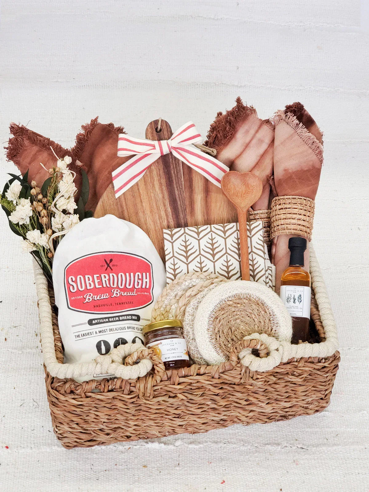 woven gift basket with sourdough bread mix, honey jar, olive oil, wooden cutting board, napkins, and kitchen utensils