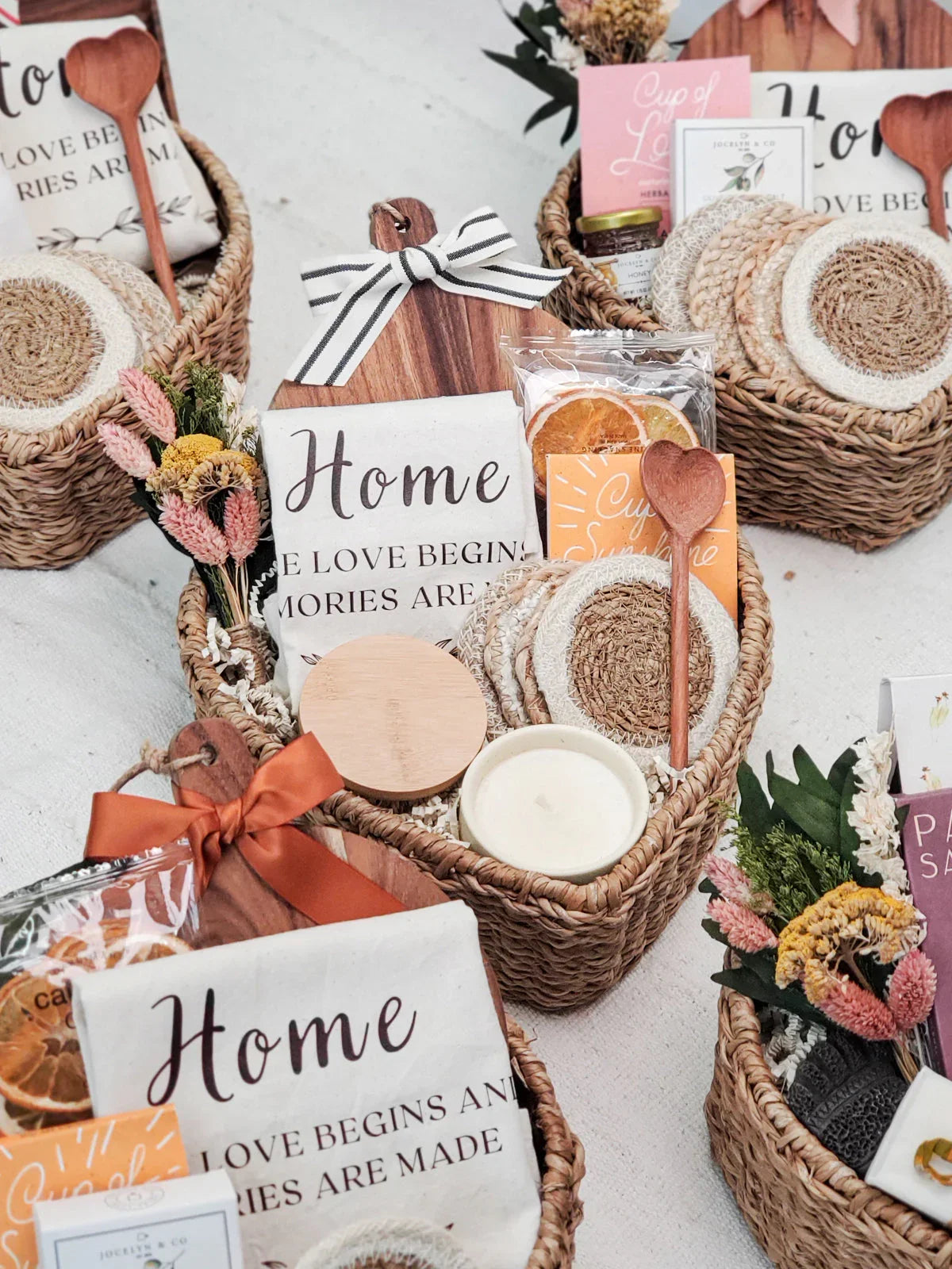 Woven gift baskets with home decor towels, heart-shaped wooden spoons, coasters, candle, dried flowers, and packaged treats