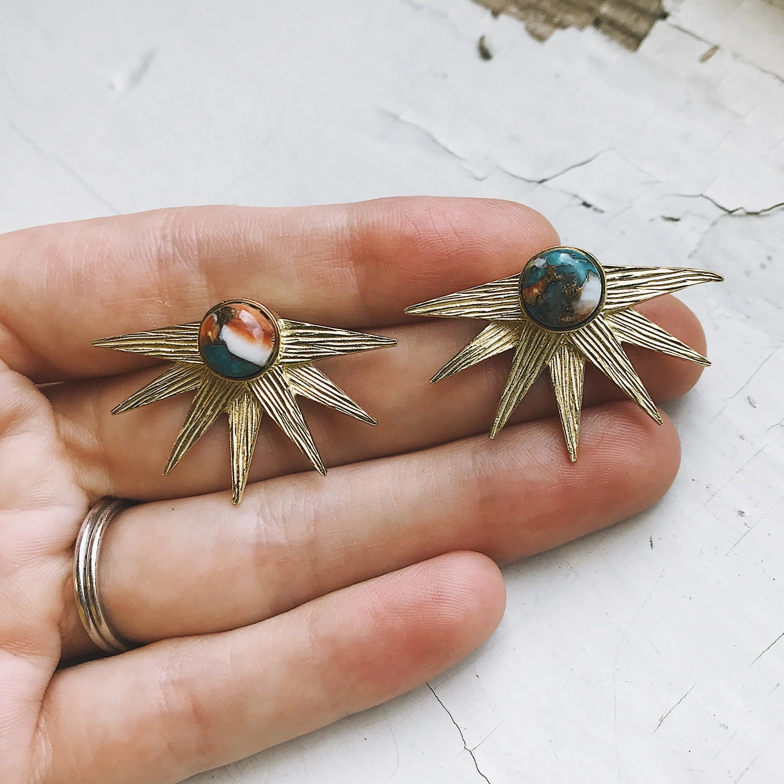 Hand holding two gold sunburst earrings with multi-color stone centers on cracked white surface
