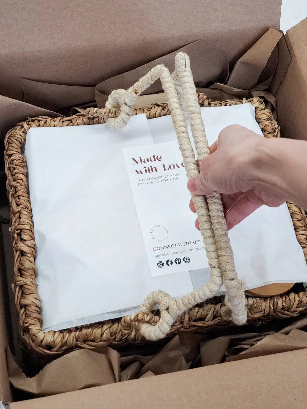 Hand holding handle of woven gift basket with 'Made with Love' note inside a cardboard box