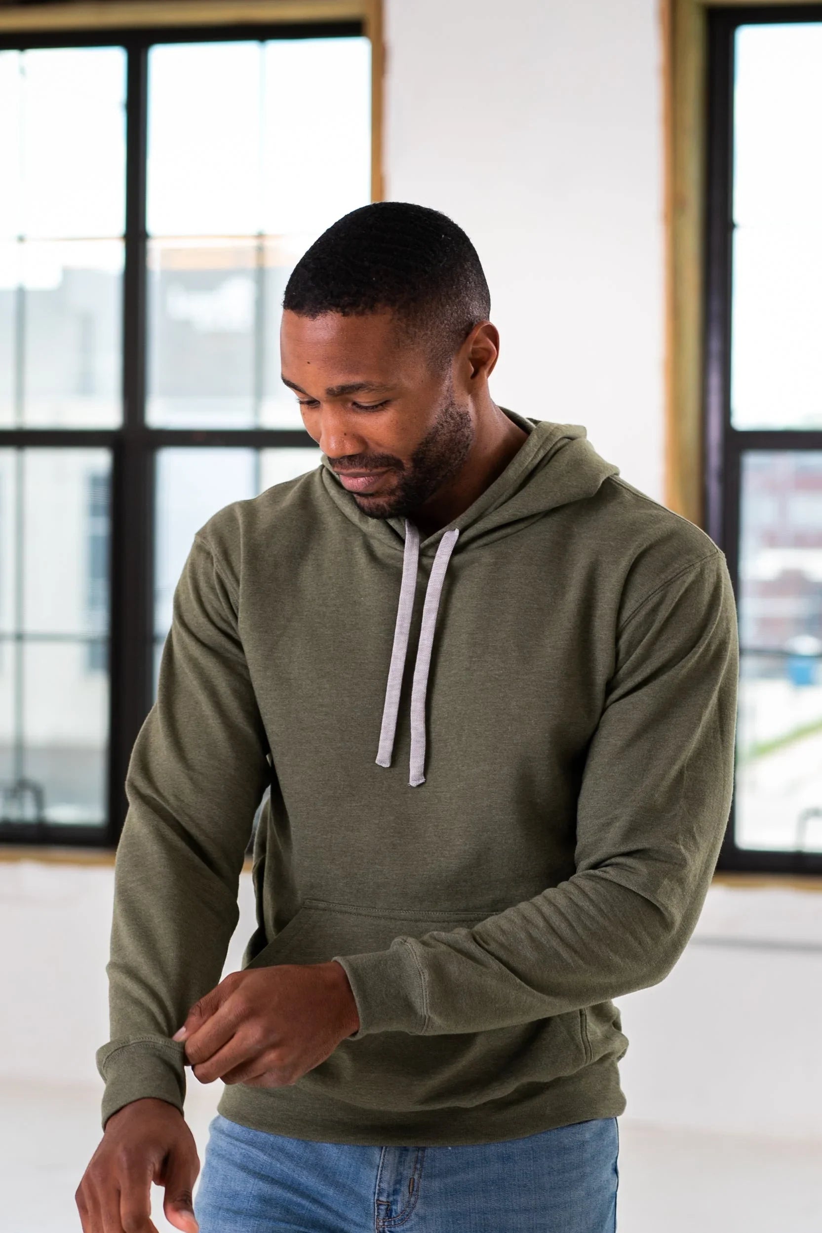 Man in olive green hoodie and jeans standing indoors near large windows at Qu!rk Boutique