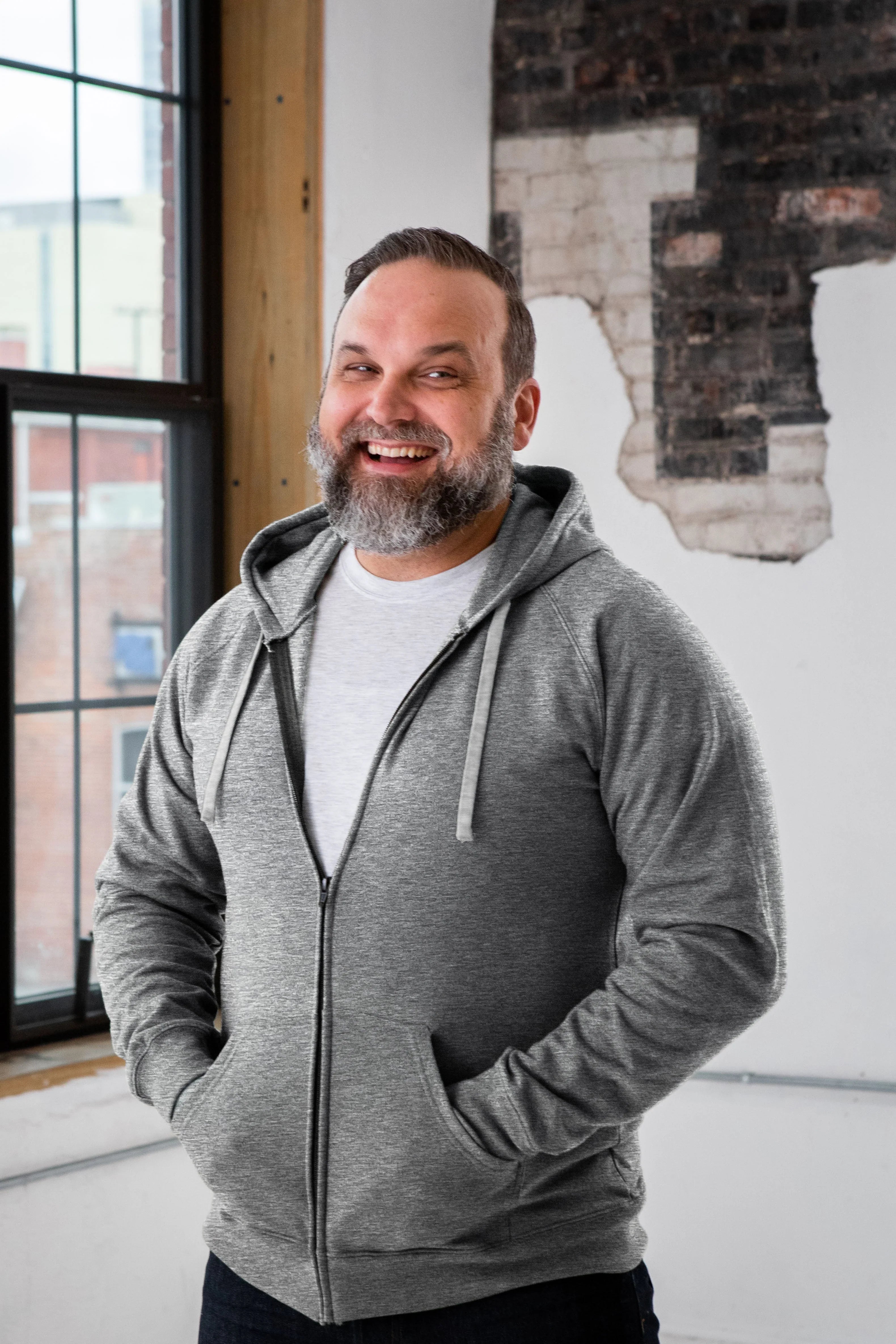 Smiling man in a gray zip-up hoodie and white t-shirt, standing indoors by a window at Qu!rk Boutique.