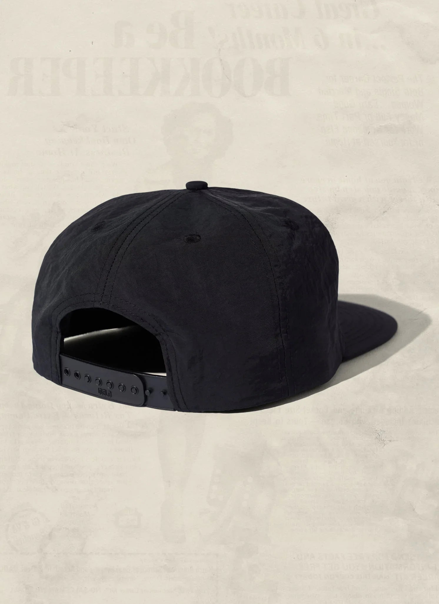 Back view of a black snapback cap from Qu!rk Boutique on a neutral textured background