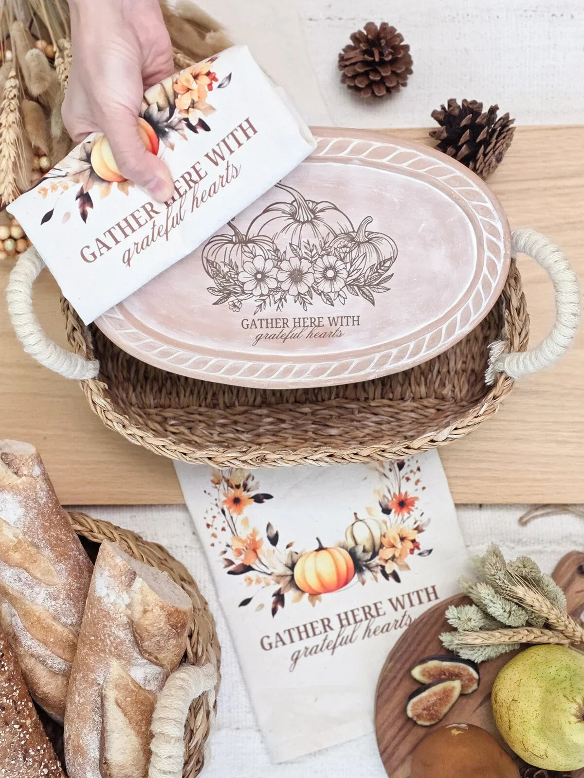 Hand holding autumn-themed cloth with "Gather here with grateful hearts" text over wicker basket and rustic fall serving tray