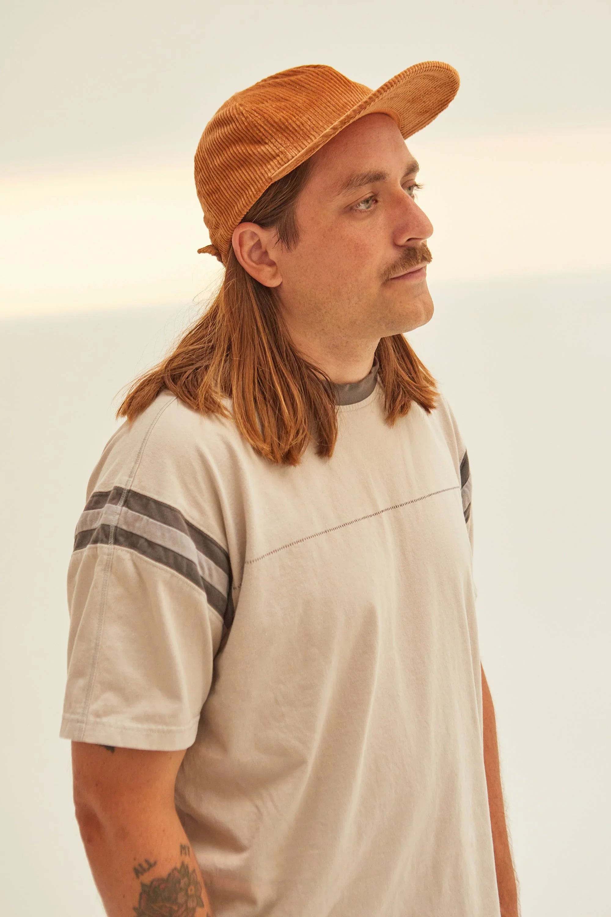 Person in tan shirt with grey stripes and brown corduroy cap, neutral background, Qu!rk Boutique style