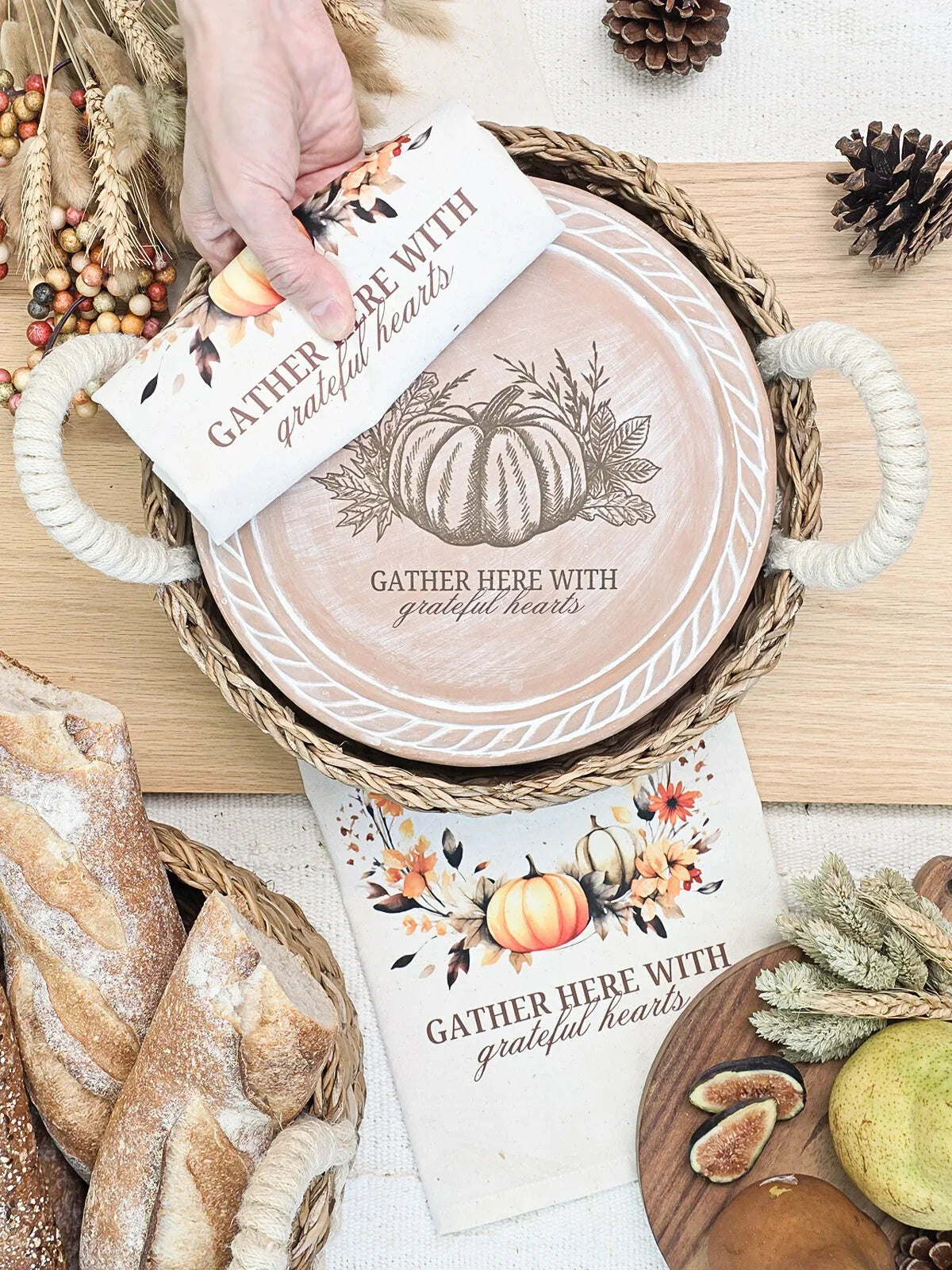 Rustic autumn kitchen scene with woven tray, bread, pears, figs, pinecones, and fall-themed towels and plate saying 'Gather here with grateful hearts'