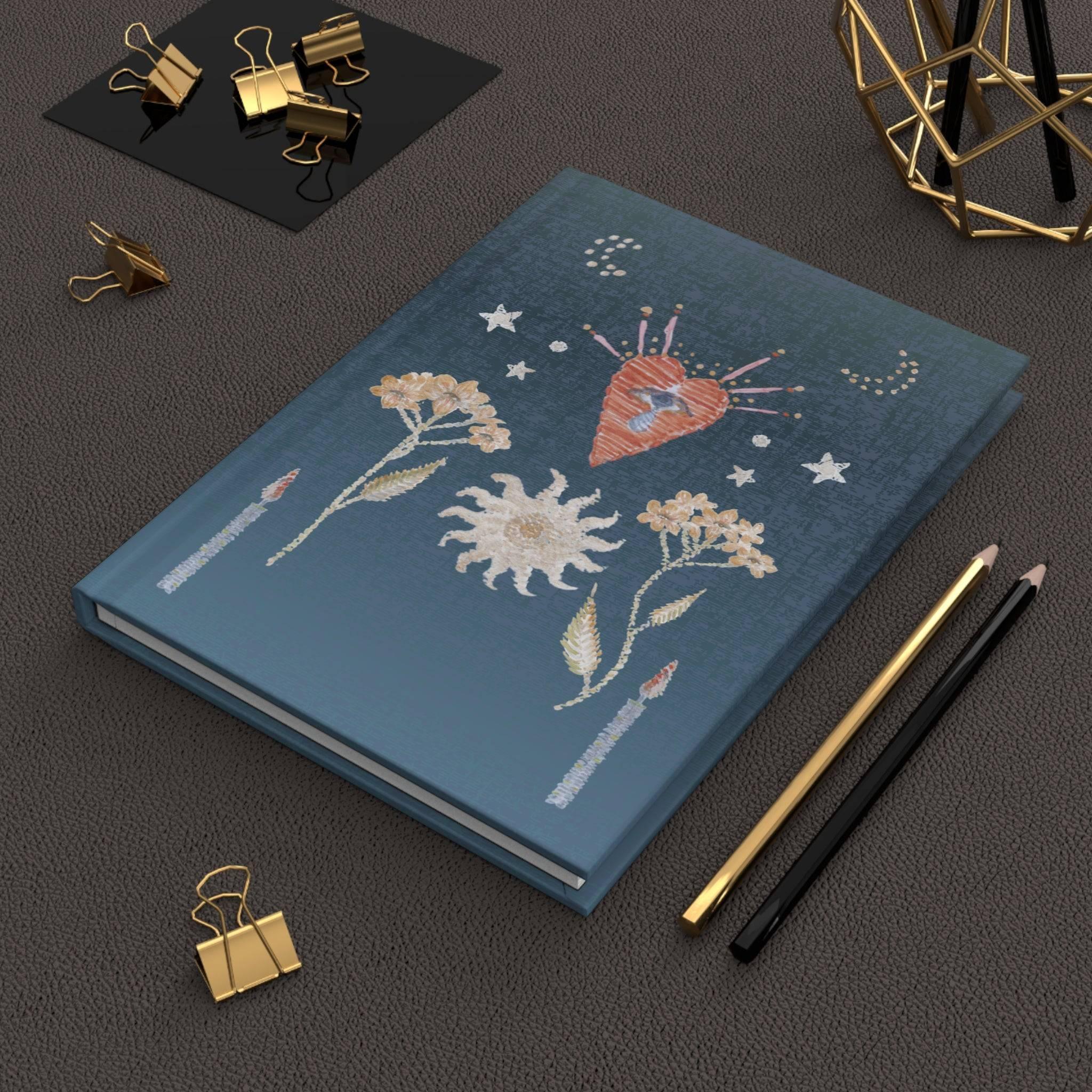 Blue hardcover notebook with embroidered floral, heart, stars, and candle designs, on a textured dark surface