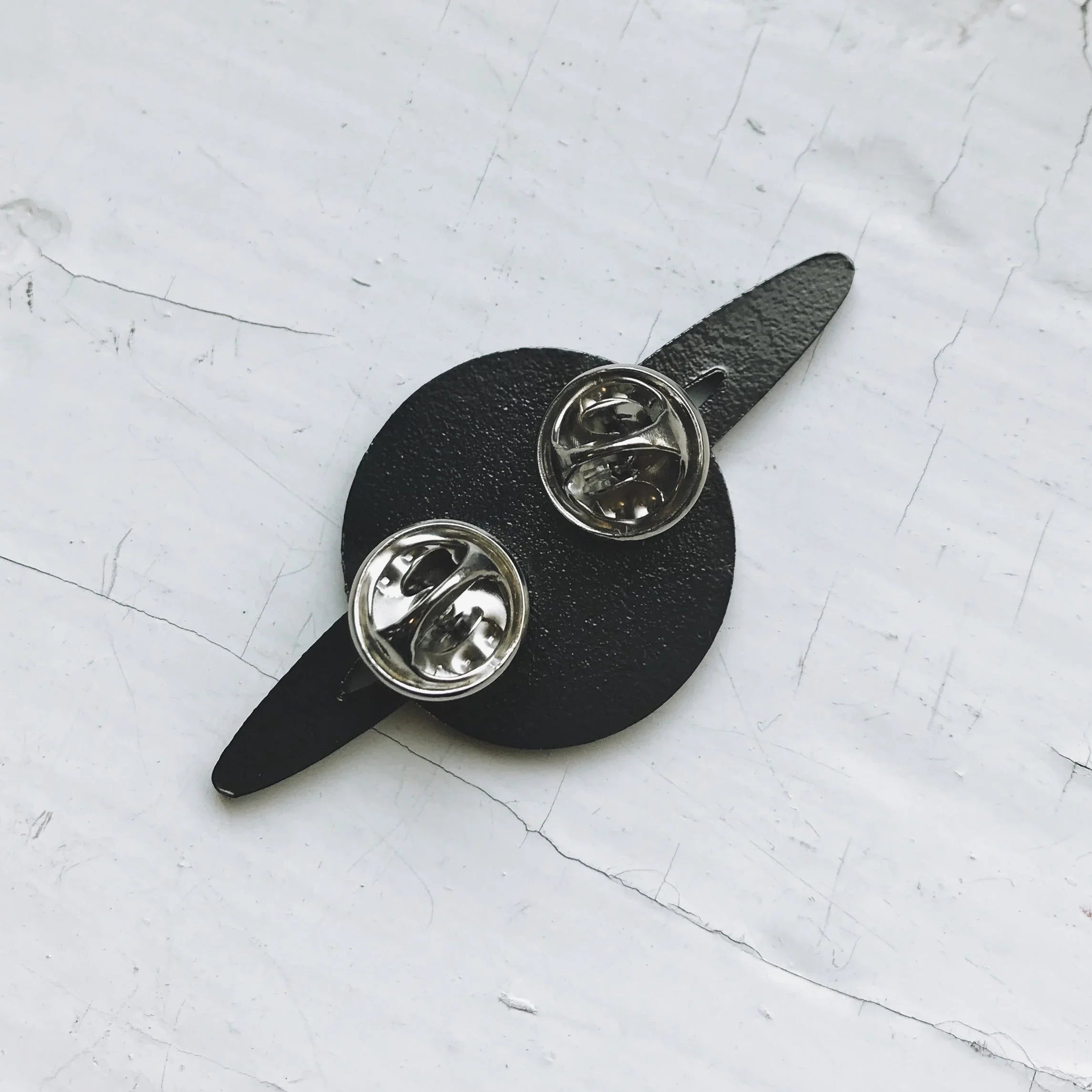Black leather pin backing with two silver butterfly clutch pins on cracked white surface